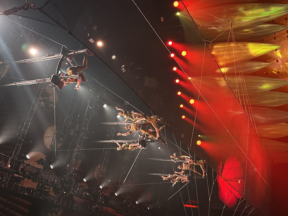 A low-angle shot shows acrobats mid-performance in a dimly lit venue. The performers are suspended from metal structures, some hanging upside down. A black net is visible above. A large screen to the right emits a red light, punctuated by rows of illuminated circles. Yellow and green petal-like shapes appear above the screen, suspended by thin white cords. The stage's metal framework and lighting equipment are partially visible.