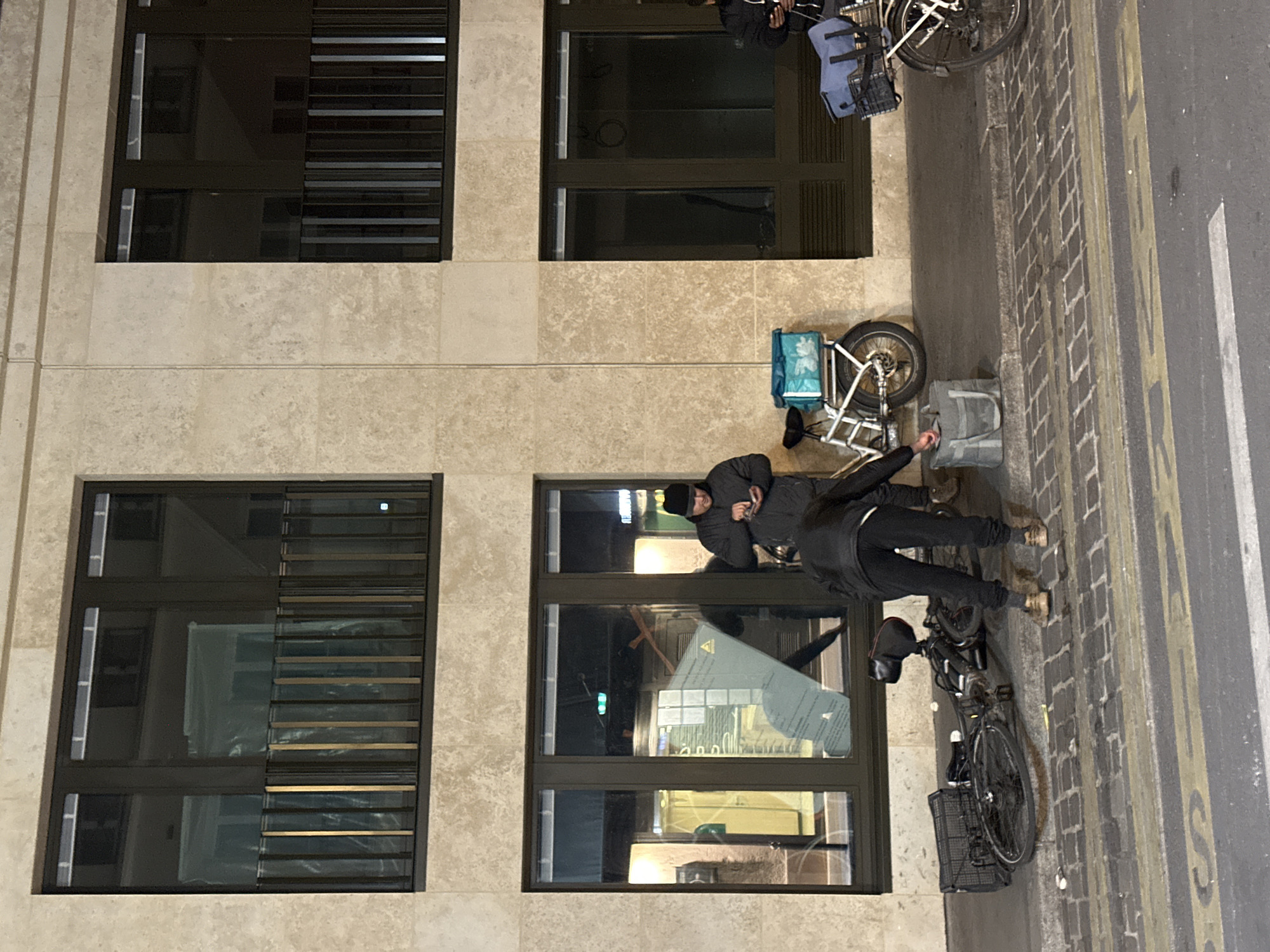 A man in a dark hooded jacket and dark pants stands on a brick-paved sidewalk next to a parked bicycle with a rear basket. The sidewalk is adjacent to a street with yellow lines and white lane markings. Behind the man, against a beige building facade, are two large, dark-framed windows, each with multiple panes. The windows reflect the man and his immediate surroundings. A second bicycle with a delivery container attached is parked on the sidewalk further away. The location is likely in a European city, potentially Paris, France, based on the architectural style and street markings.