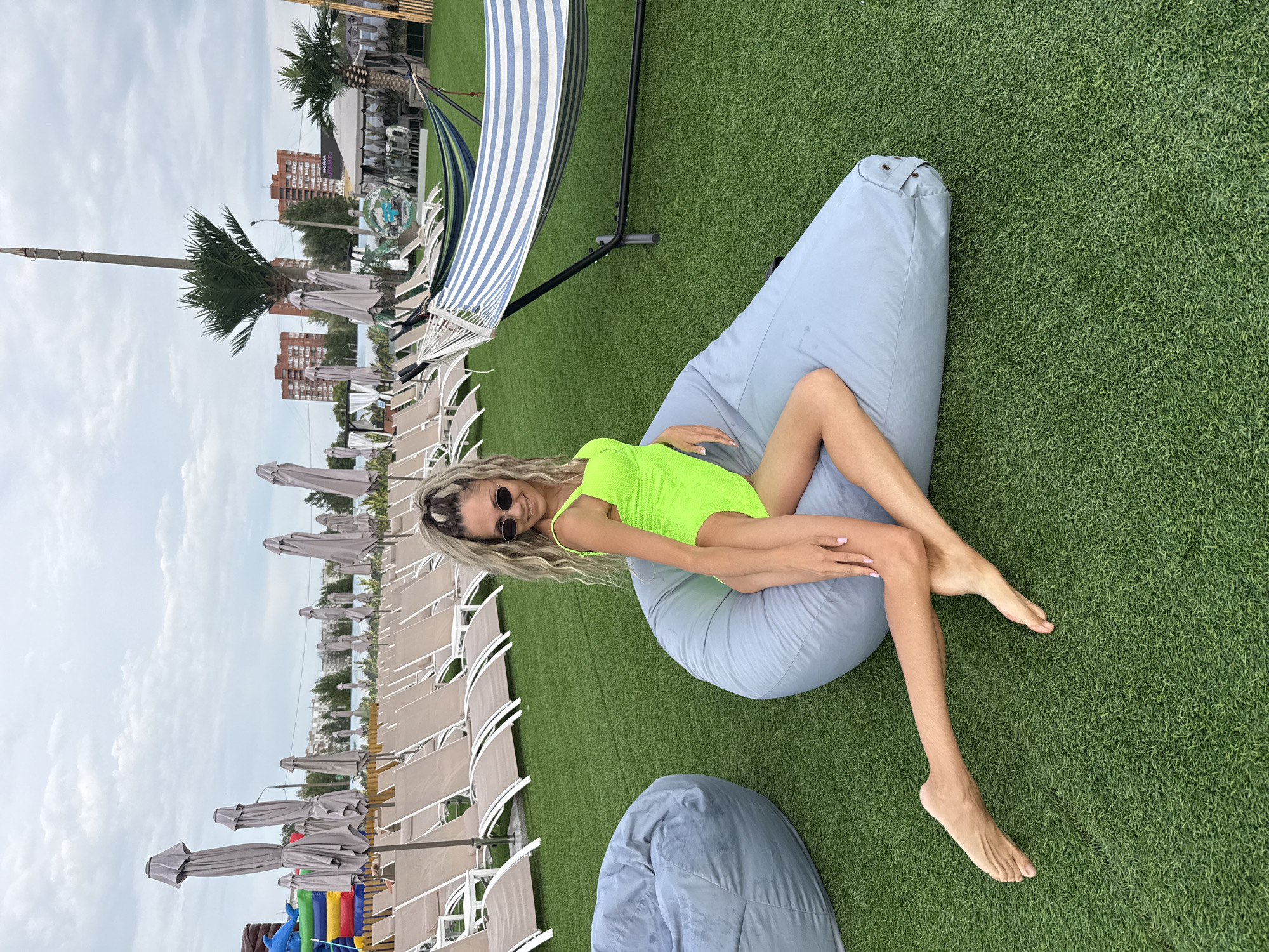 A woman in a neon green swimsuit is sitting on a large, light blue beanbag chair on a patch of artificial turf. She is positioned in the foreground, facing the right side of the frame, with her legs crossed and extended. Her left leg is bent at the knee, with her foot resting on the beanbag, while her right leg is extended forward. She has blonde, wavy hair and is wearing oval sunglasses. In the background, rows of empty lounge chairs are visible, along with closed umbrellas. Further in the distance, hammocks are set up, and buildings and trees can be seen under a cloudy sky. The location is identified as a resort in Rusanovo, Russia.