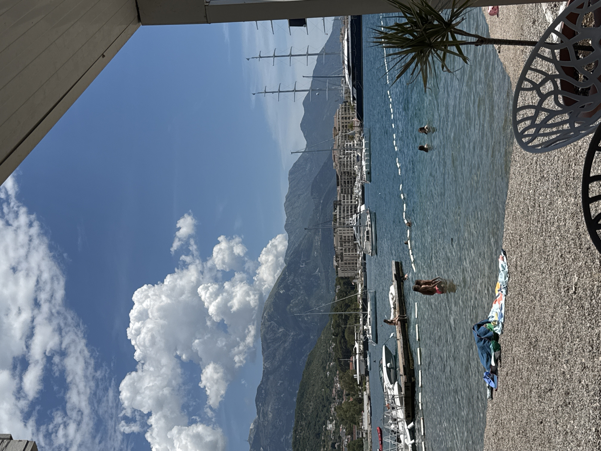 The image depicts a scenic view of a marina in Tivat, Montenegro, on a bright, sunny day with scattered clouds. The foreground shows a pebble beach with towels and beach gear laid out, suggesting a recreational area. In the mid-ground, the calm blue water of the bay is dotted with various boats, including several sailboats and a very large, dark-hulled yacht with multiple masts. A wooden pier extends into the water, where a few people are visible, some sitting on the edge and others wading in the shallow water. Further out in the bay, more vessels are anchored or moored. In the background, lush green hills rise, forming a backdrop to the marina, and beyond them, majestic mountains with rocky peaks are silhouetted against the clear sky. Part of a modern building structure is visible in the right foreground, framing the view and suggesting a waterfront establishment.