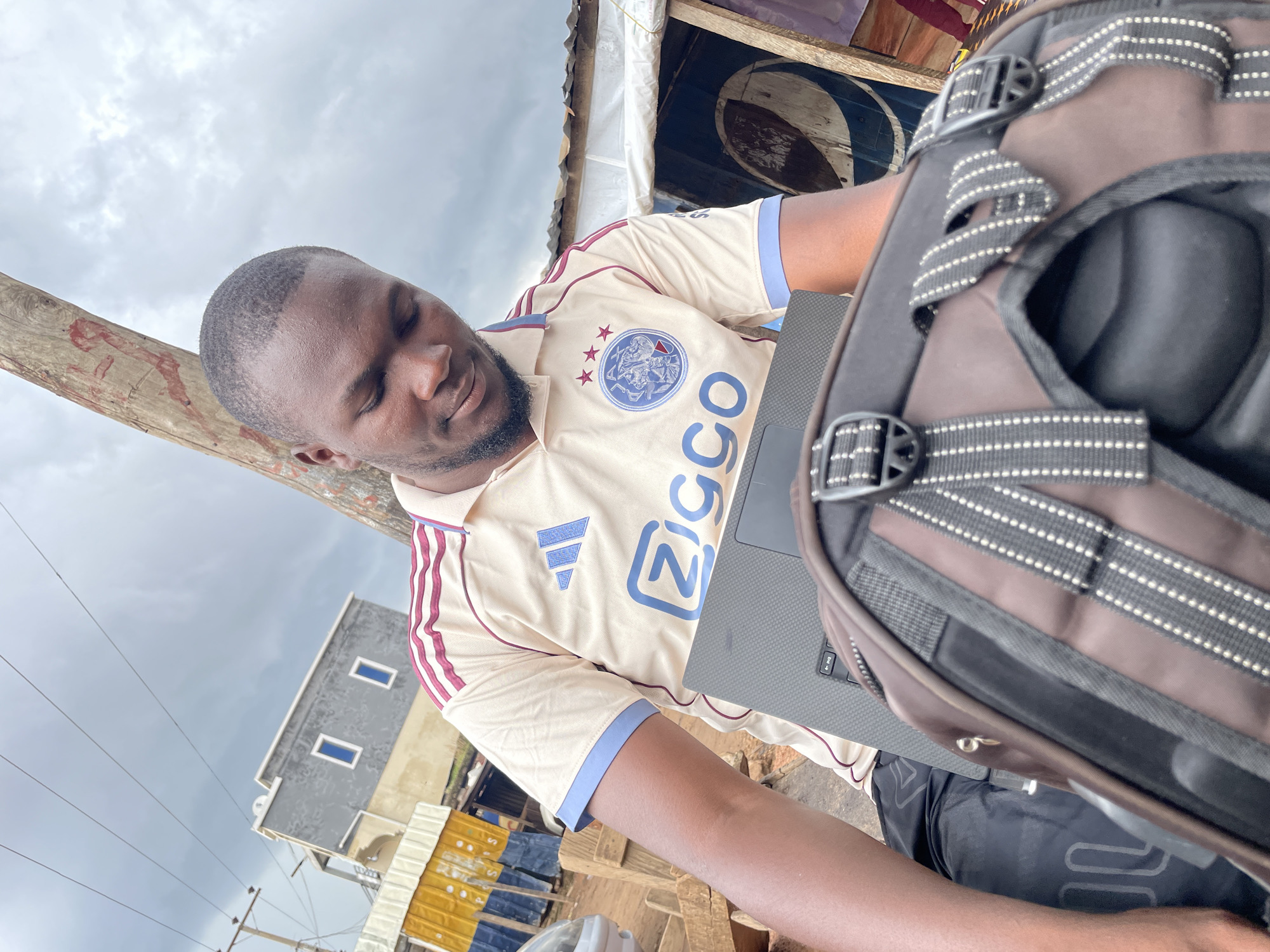This is a portrait of a Black man in Jos, Nigeria, seated outdoors and focused on a laptop. He is wearing a cream-colored polo shirt with maroon and blue trim, featuring an Adidas logo and what appears to be the Ajax football club crest. The man has his eyes closed, suggesting a moment of concentration or rest, and has a beard.
In the foreground, a portion of a dark brown backpack with reflective stripe detailing is visible, positioned in front of the man and partially obscuring the laptop. The laptop's keyboard is visible, indicating it is open and in use.
The background suggests an outdoor, somewhat informal setting. A wooden pole is to the left of the man. Beyond that, there are hints of a stall or shop with a Pepsi logo visible on a sign. Further in the distance, a two-story building with a grey and cream facade and visible windows can be seen, along with power lines and other structures that suggest an urban or semi-urban environment. The sky is overcast with clouds, indicating a cloudy day. The overall mood is one of quiet focus amidst an everyday setting. There is visible text on the man's shirt: the Adidas logo, the Ajax crest, and the word "Ziggo" is partially visible. The Pepsi logo is also visible in the background.