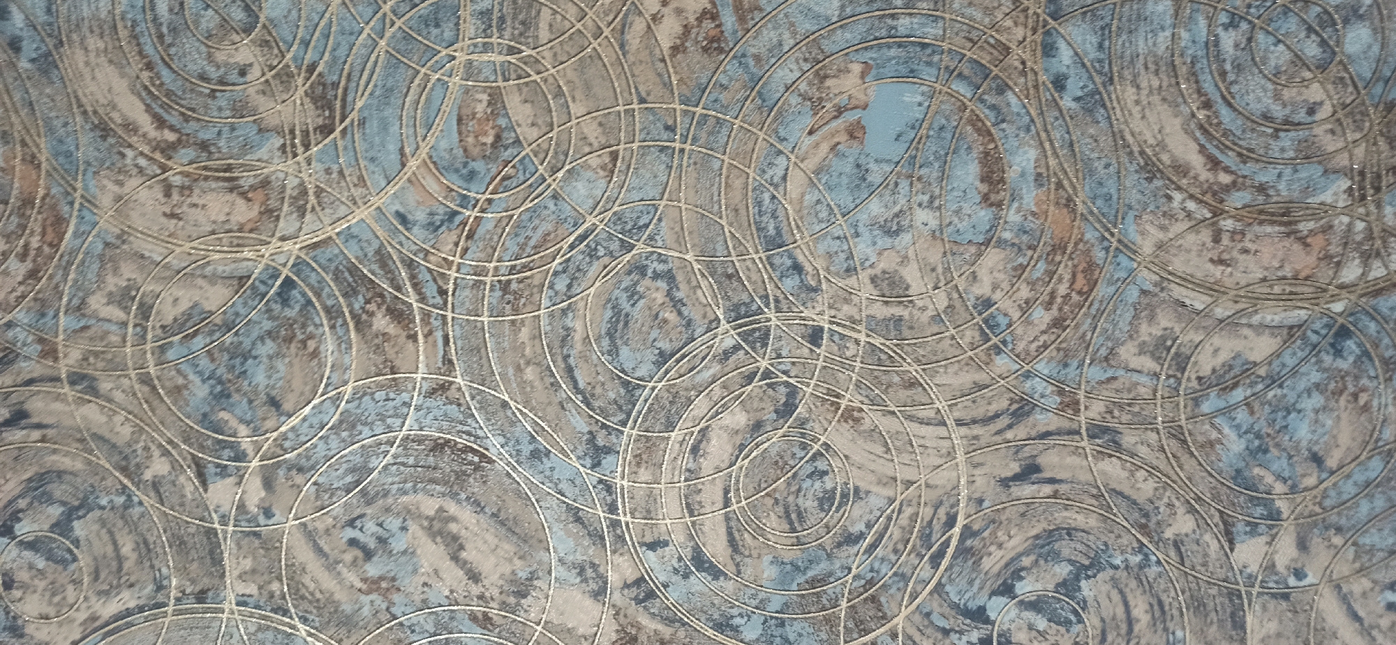 The image displays a close-up view of a textured surface, likely wallpaper or fabric, featuring an abstract pattern of overlapping circles and swirls. The colors are predominantly muted blues, grays, and earthy browns, with thin, shimmering silver lines defining the circular motifs. The texture appears somewhat rough or distressed, with variations in color and shading suggesting a layered or marbled effect. There are no discernible people, objects, or scenes. The lighting is even, with no indication of time of day or weather. The location context of Asbest, Russia, does not provide any visual cues within the image itself. No text is visible.