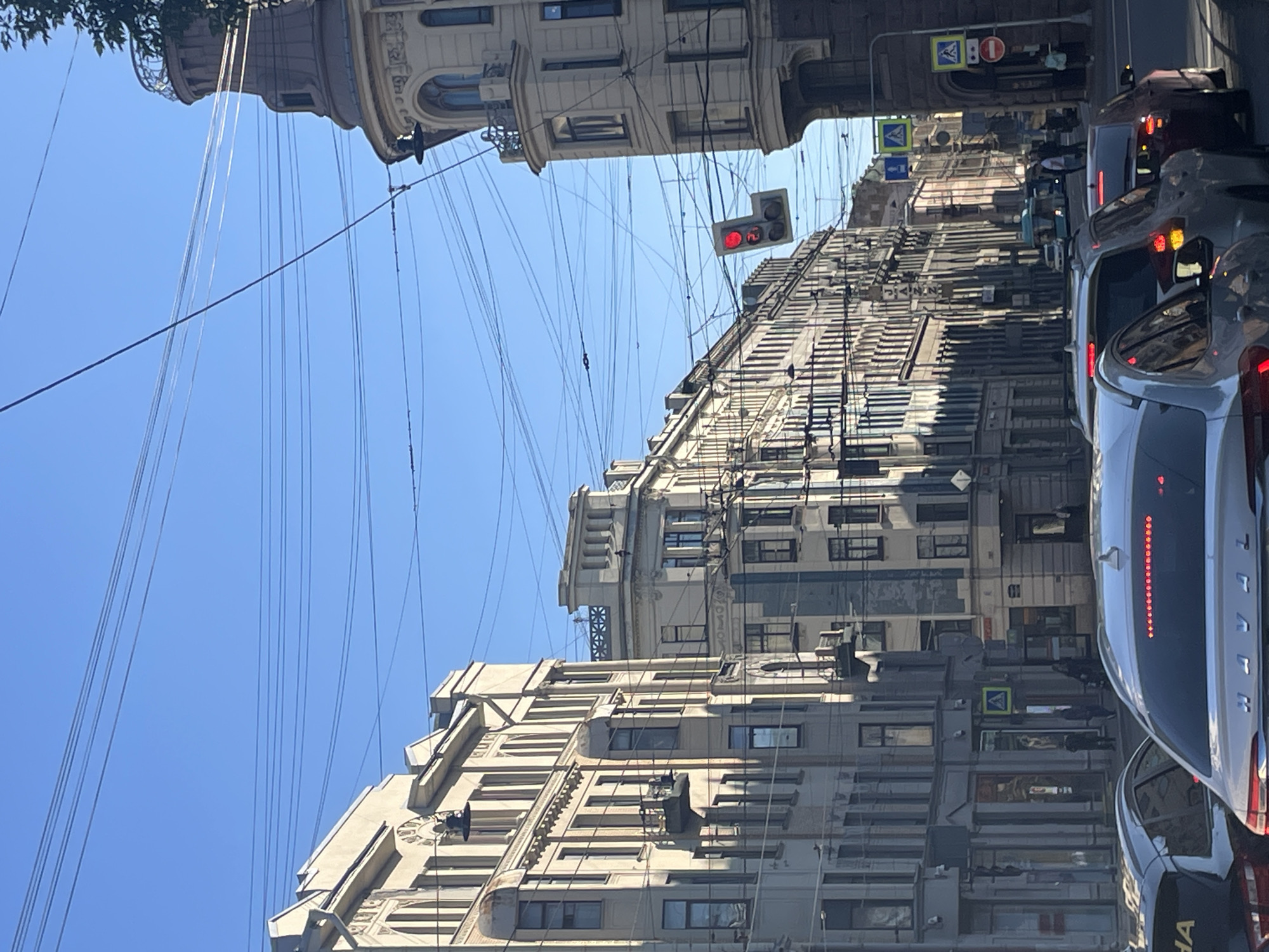 The image captures a sunlit street scene in Saint Petersburg, Russia. The architecture is characterized by ornate, multi-story buildings with classical detailing, indicative of the city's historical aesthetic. Several cars are stopped in traffic, suggesting a busy urban environment. Overhead, a complex network of power lines and streetlights crisscrosses the sky, a common feature in older city districts. A traffic light is visible, displaying a red signal. Pedestrians are seen on the sidewalks, and signs of various types, including directional and traffic signals, are present. The clear blue sky suggests fair weather. The buildings on the left are lighter in color, while those on the right appear to be of a darker hue, with a prominent domed tower structure visible. The overall impression is of a well-established European city street with a blend of historical architecture and modern urban activity.