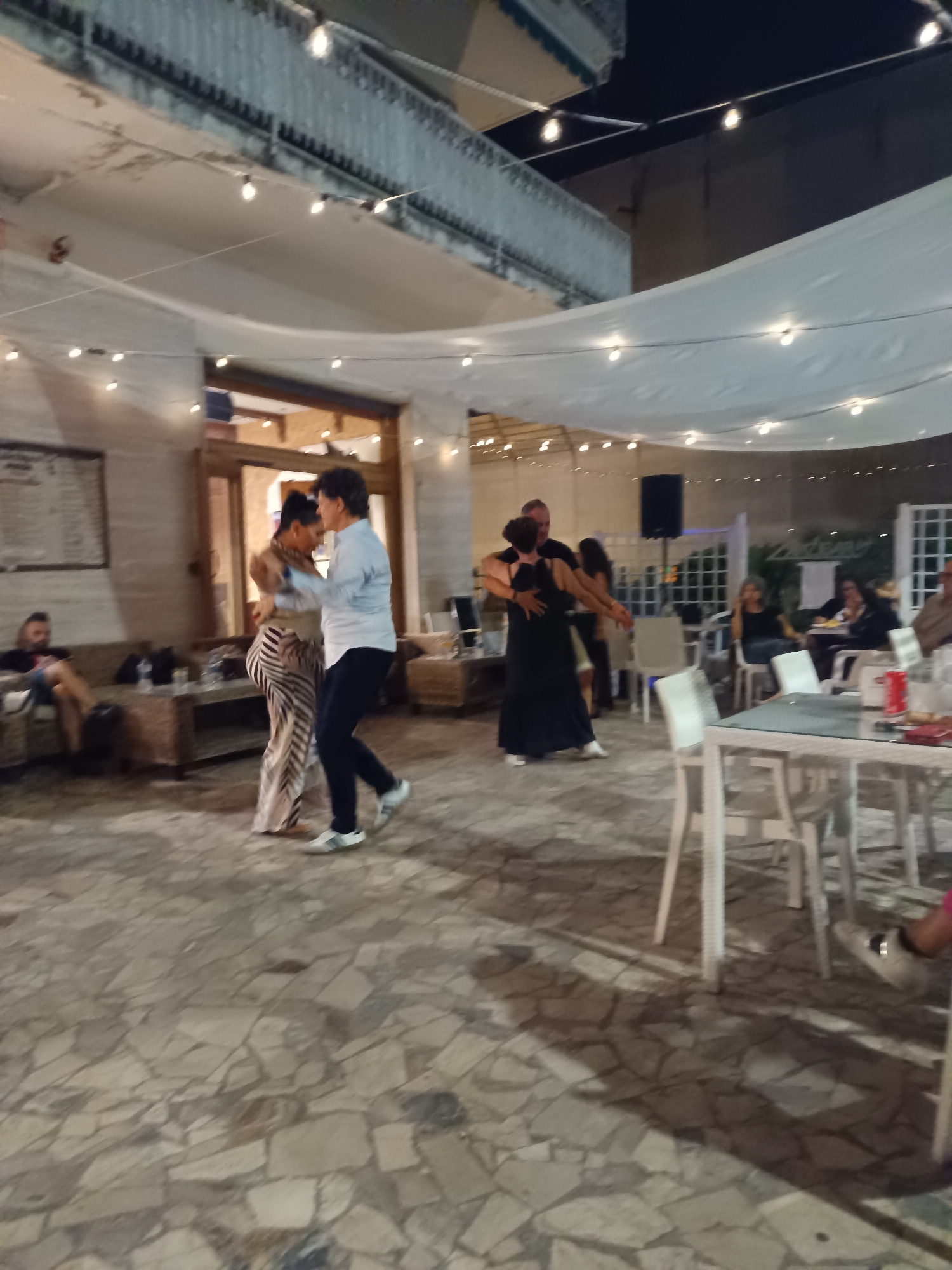 The image depicts an evening scene at an outdoor or semi-outdoor venue in Pineto, Italy, characterized by social activity. The ground is paved with irregular light-colored stone slabs. Overhead, numerous string lights illuminate the area, alongside white fabric drapes suspended across the space.
In the mid-ground, two couples are actively engaged in partner dancing. To the left, a woman in a striped (light and dark) wide-leg pantsuit and a man in a light blue button-up shirt and dark pants are dancing, facing each other. To the right, another couple, a woman in a dark dress and a man in a dark shirt and pants, are also dancing, holding each other in a standard partner dance pose.
On the left side of the scene, a man with dark hair and a beard is seated on a wicker-style sofa, looking towards the right, wearing a dark t-shirt and light shorts. Adjacent to him are low tables. In the background on the left, a building with a large wooden double door entrance is visible, and a wall features a menu board or sign.
The right side of the image features multiple white tables and chairs, some occupied by individuals. One woman with light hair is seated, wearing a dark top. Other figures are visible further back, appearing to be dining or socializing. A large black speaker on a stand is positioned in the mid-right background. White latticework structures are also present in the background on the right. The upper background reveals a dark sky and elements of a building structure or balcony. The overall ambiance is lively and social, consistent with an evening event at a restaurant or resort.
