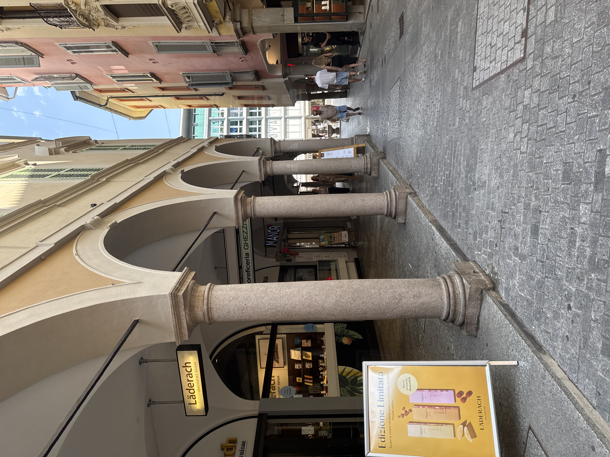 The image depicts a narrow cobblestone street in Lugano, Switzerland, lined with arcades and buildings of varying colors. A prominent sign for "Läderach chocolatier suisse" is visible, along with a display for their limited edition products in front of the store. Several people are walking down the street, some appearing to be tourists. The architecture suggests a European city center.