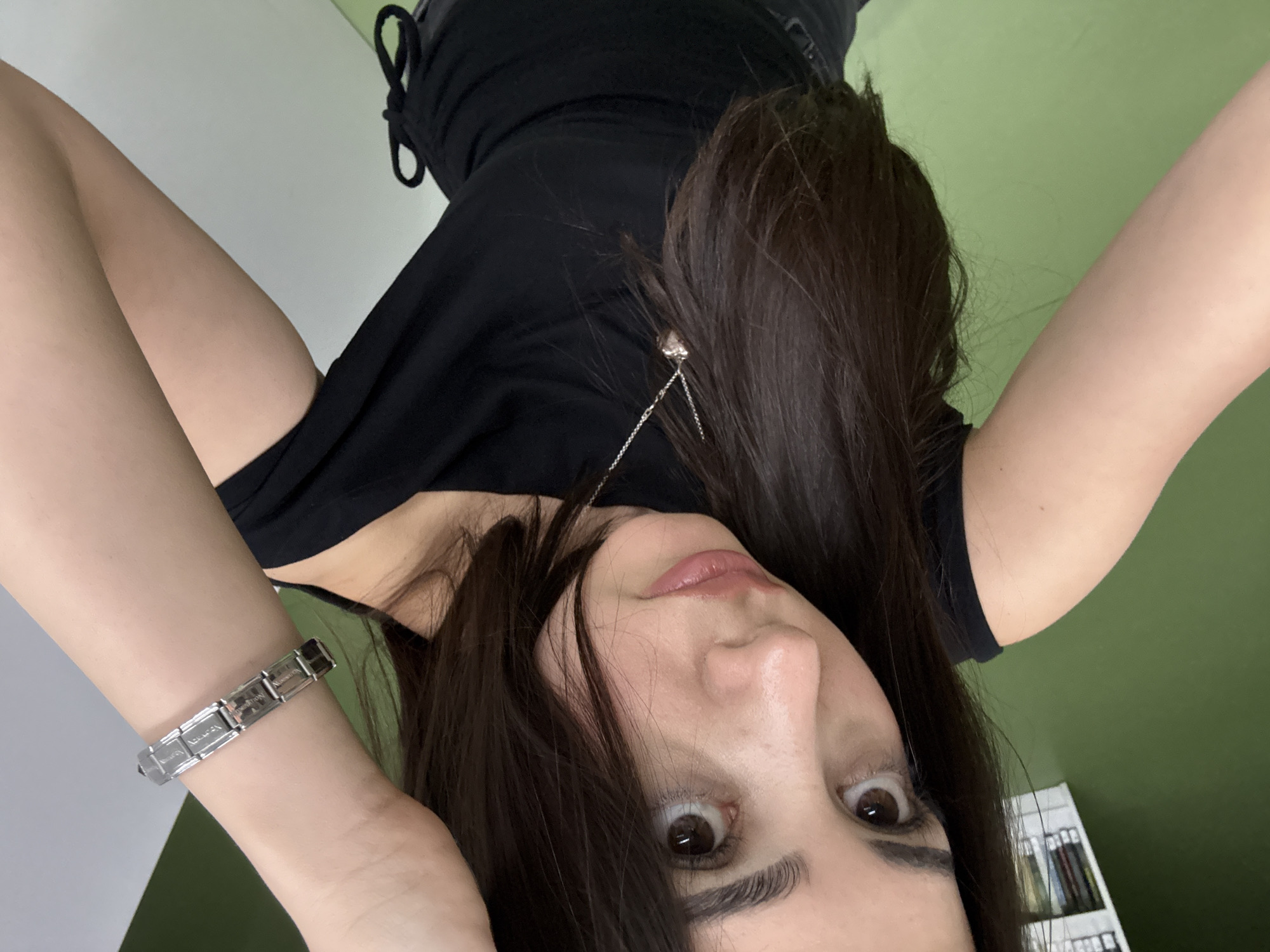 A female individual is depicted in a close-up selfie, captured indoors in Yerevan, Armenia. She has long, dark brown hair and is wearing a black off-the-shoulder top with a visible gathered tie on the lower right side. A silver-colored chain necklace with a small heart-shaped pendant is visible on her chest. Her right hand is raised, with her wrist adorned by a silver-colored link bracelet, identifiable as a modular type. Her left arm is visible to the left. The individual has dark eyes and defined eyebrows, gazing upwards and slightly past the camera, with a neutral to slightly pouty expression. The background consists of a solid green wall on the left, with a partial view of a white shelf unit containing books or binders in the upper left corner. A plain white wall or vertical surface is visible on the right side of the frame.
