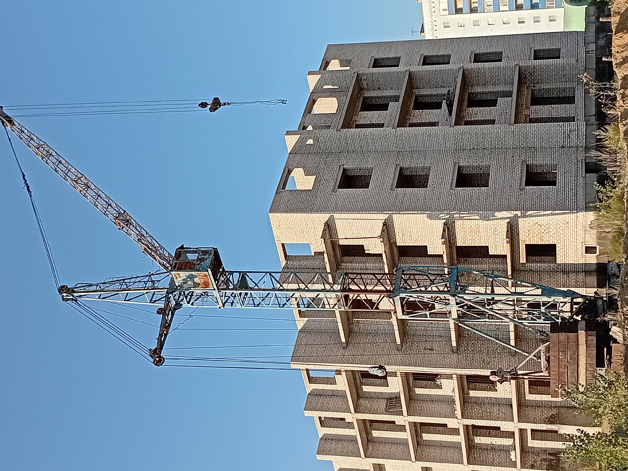 The image shows a construction site with an unfinished brick building and a tower crane. The building is multi-story, with exposed brickwork and open window frames. The tower crane is a prominent feature, with its boom extending over the building. A hook hangs from the crane's cable. The sky is clear and blue. In the background, a partially visible, completed apartment building can be seen. The scene appears to be in the city of Semey, Kazakhstan, a location often associated with such construction projects as the region undergoes development. The overall impression is one of ongoing construction work under a clear sky.