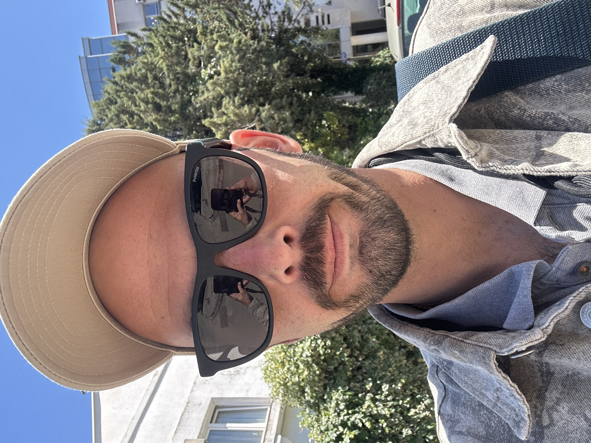 A selfie taken by a man with a beard, wearing sunglasses and a hat. The reflection in the sunglasses shows him taking the picture with a phone. He is wearing a light denim jacket over a grey shirt, with a blue strap visible on his shoulder. The background shows trees, a building, and a clear blue sky. The location is likely Budva, Montenegro, based on building architecture and foliage.