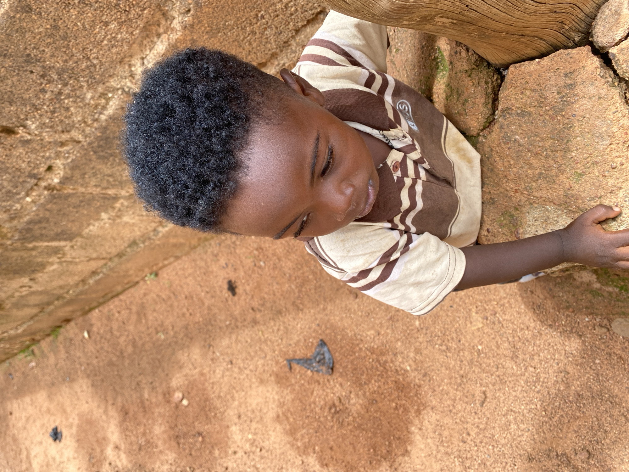 The image, taken in Kuru Dungus, Nigeria, presents a high-angle, close-up view of a young boy in an outdoor setting. The boy has dark skin and short, dense, black curly hair. He is looking downwards, with his eyes partially closed or cast low, and his mouth slightly ajar, conveying a gentle or contemplative expression. He is wearing a short-sleeved collared shirt with vertical stripes of alternating light beige/cream and brown. A white and gray design, resembling a stylized spiral or an 'S' shape, is visible on the upper left chest area of his shirt.
He is positioned alongside a rustic environment, with his left arm extended, and his hand resting on or near large, rough, reddish-brown rocks. Some of these rocks show hints of green moss. To his right and behind him is a large, weathered wooden beam or post. The ground beneath him is a reddish-brown, earthy, or sandy surface. A small, dark, irregular piece of debris lies on the ground in the foreground. The scene is illuminated by natural daylight, casting warm tones over the surroundings.