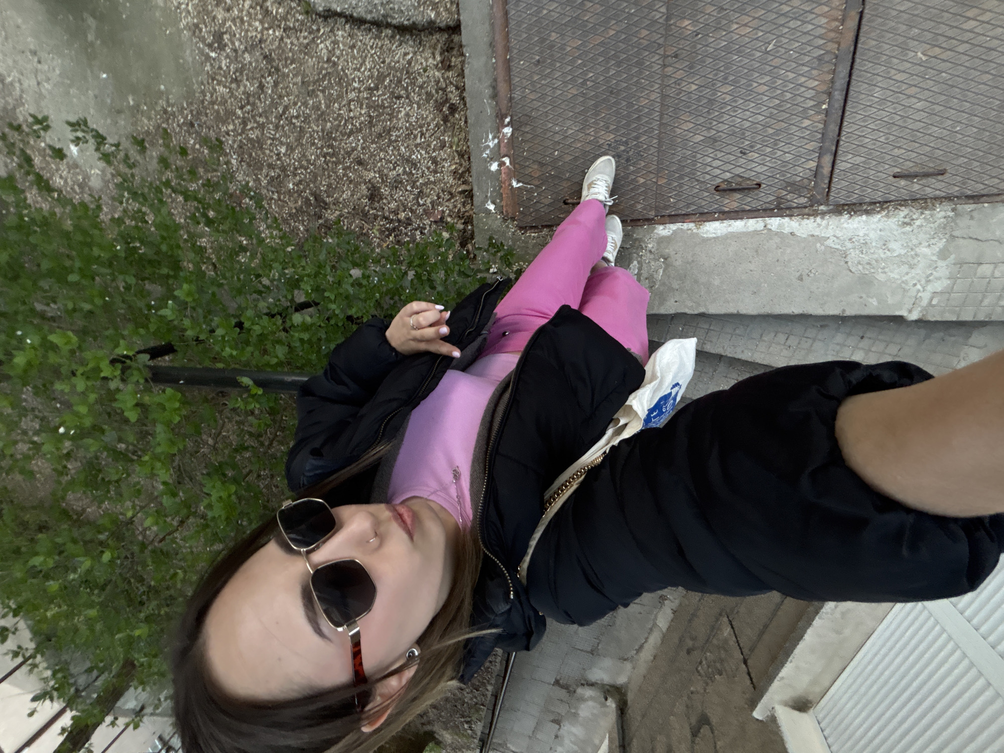 A person stands on a concrete surface adjacent to a metal grate. The person is wearing a black jacket, pink pants, white shoes, and sunglasses. A bush with green leaves is visible on the left. The surrounding area appears to be an urban environment. The exact location in Veliko Tarnovo, Bulgaria cannot be identified from the information in this image alone.
