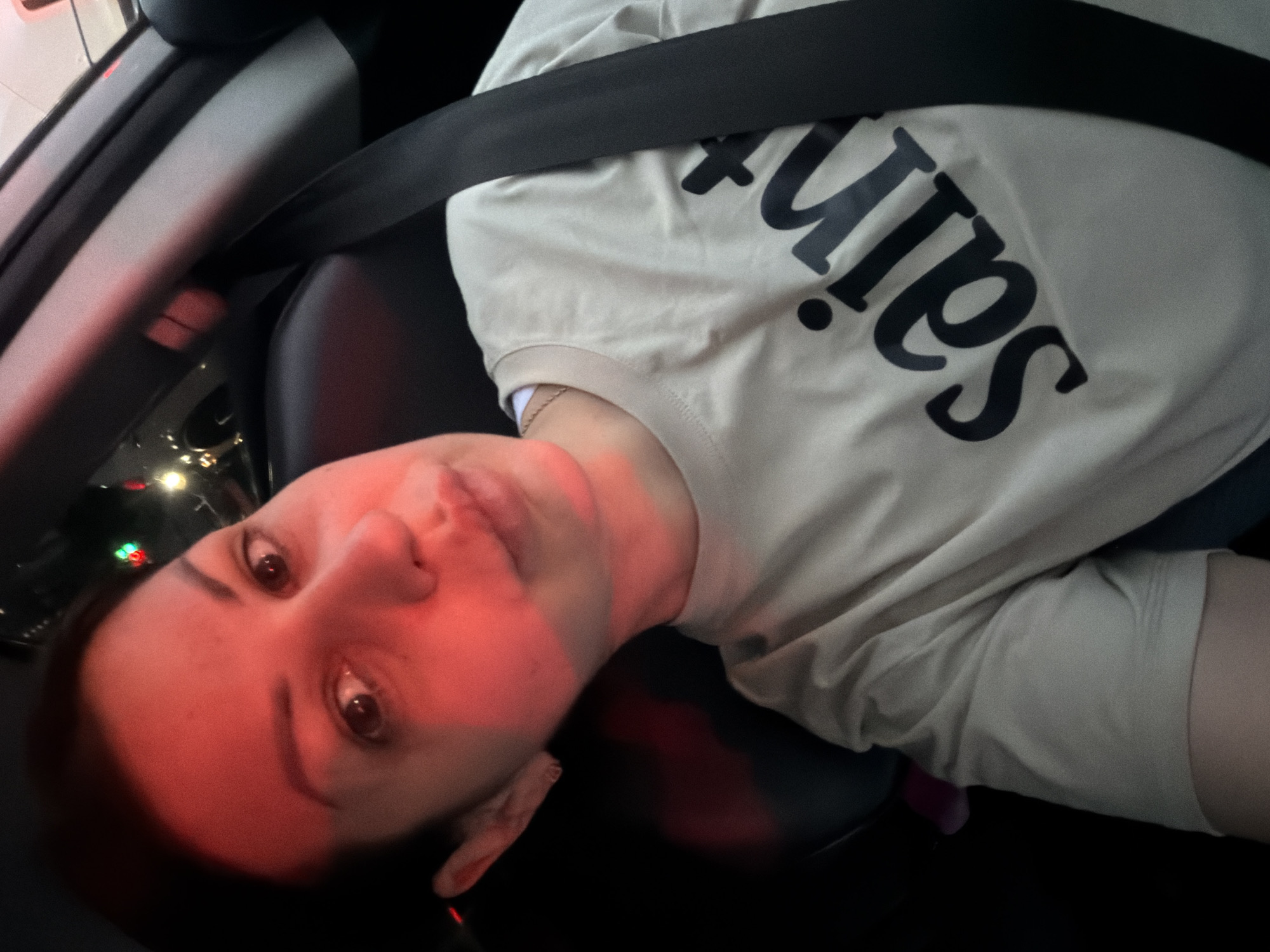 The image shows a close-up, selfie-style shot of a woman sitting in a car. She is wearing a light-colored t-shirt with the word "saint" printed in black. The car's seatbelt is visible across her chest. Red light is reflecting on her face and hair, suggesting the presence of red lighting within or outside the vehicle. The background is dark, indicating it might be nighttime or a dimly lit interior. There are blurred colorful lights visible in the upper right portion of the image, which could be traffic lights or decorative lights, potentially indicating the car is stationary in traffic or near a brightly lit area. The overall scene is focused on the woman's face and upper body. The location is not discernible from the visual information in the image.