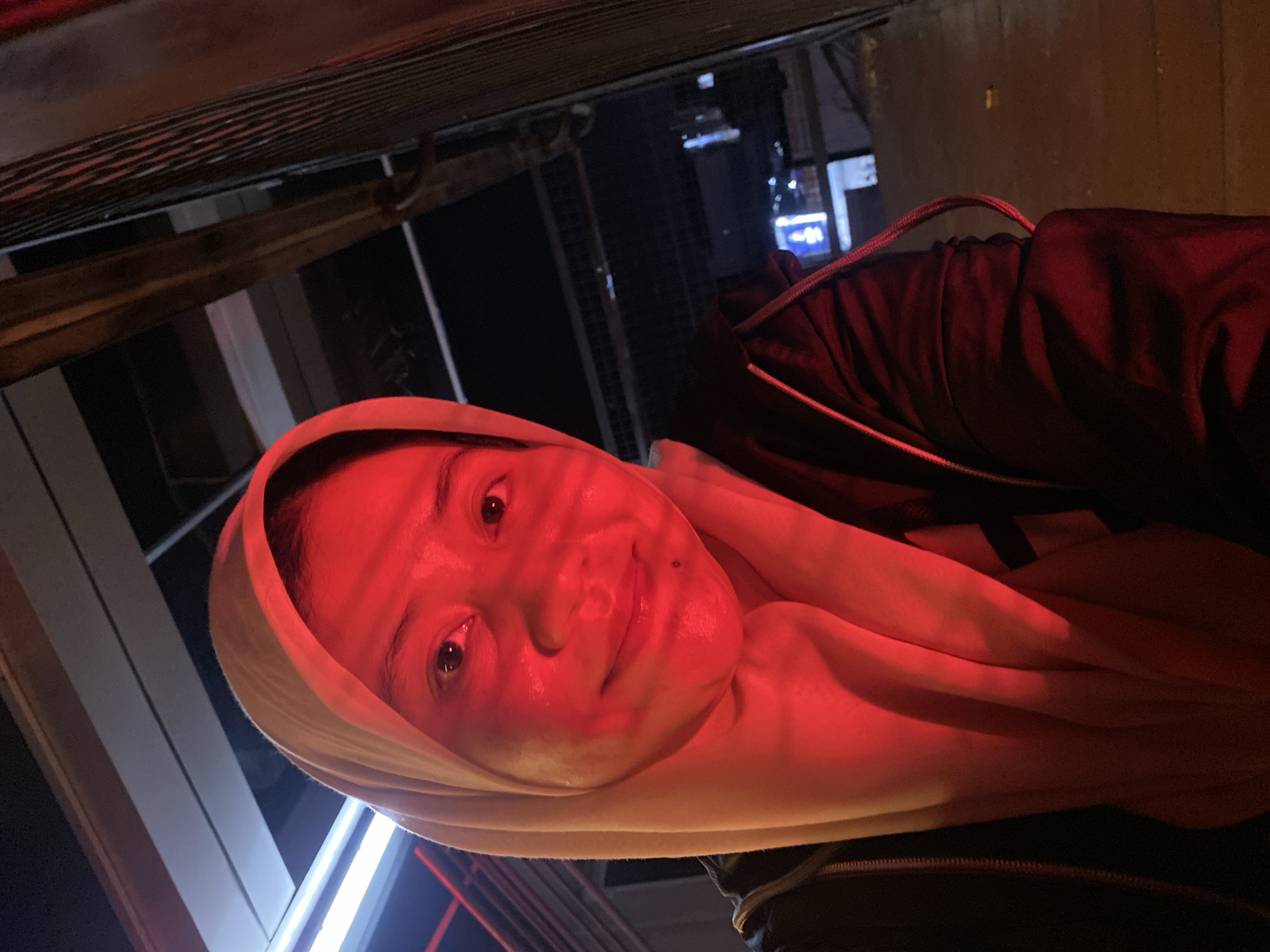 This media file depicts a close-up, low-angle shot of a person wearing a light-colored headscarf and a dark jacket. The scene is bathed in a strong red light, which casts dramatic shadows and highlights on the person's face. The person's eyes are open and looking towards the camera, with a slight smile on their lips, suggesting a relaxed or perhaps slightly playful mood. Their skin appears flushed due to the red lighting.
In the background, hints of an urban setting can be seen. There are blurred structures and lights, suggesting an outdoor environment, possibly at night or in a dimly lit area. The overall lighting and the presence of what appear to be building elements and outdoor lighting suggest an urban environment, potentially a street or an area with some form of overhead structure, like a walkway or a canopy. There are no discernible signs or text visible in the image. The time of day appears to be evening or night, given the dark background and artificial lighting. The weather is not discernible from the image.