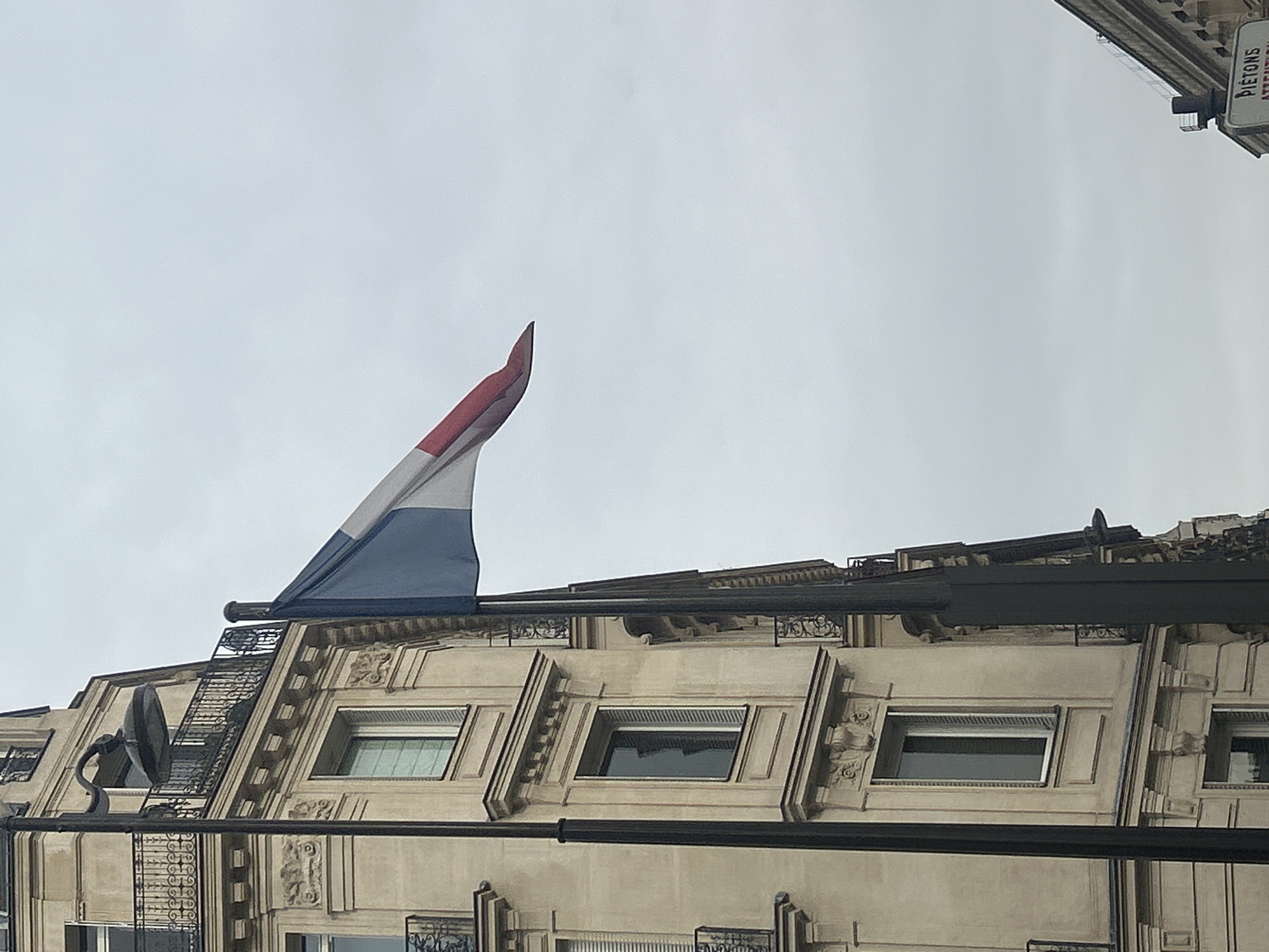 The image captures the facade of a building with architectural ornamentation. The building has multiple windows, each set within decorative frames. Above the building, a flag with red, white, and blue colors is unfurled. There is a street sign visible at the right edge of the frame.