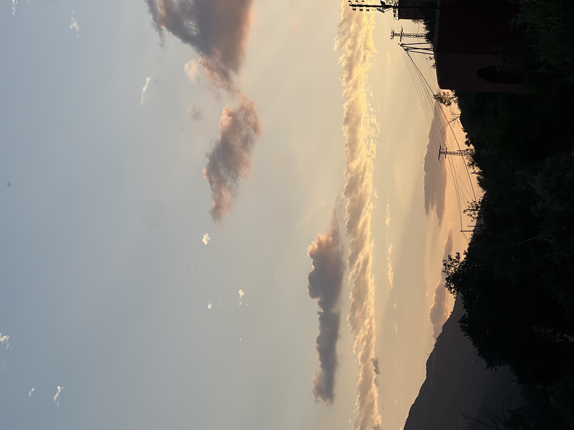 The image depicts a view of Bin El Ouidane, Morocco, at dusk. The sky is filled with long streaks of clouds with reddish hues in the distance. There are electrical poles with connecting wires and a silhouette of a mountain ridge to the bottom-right. Some dark-colored, nondescript building can be seen to the right of the electrical poles. A dense stand of trees and vegetation occupies the lower right corner.