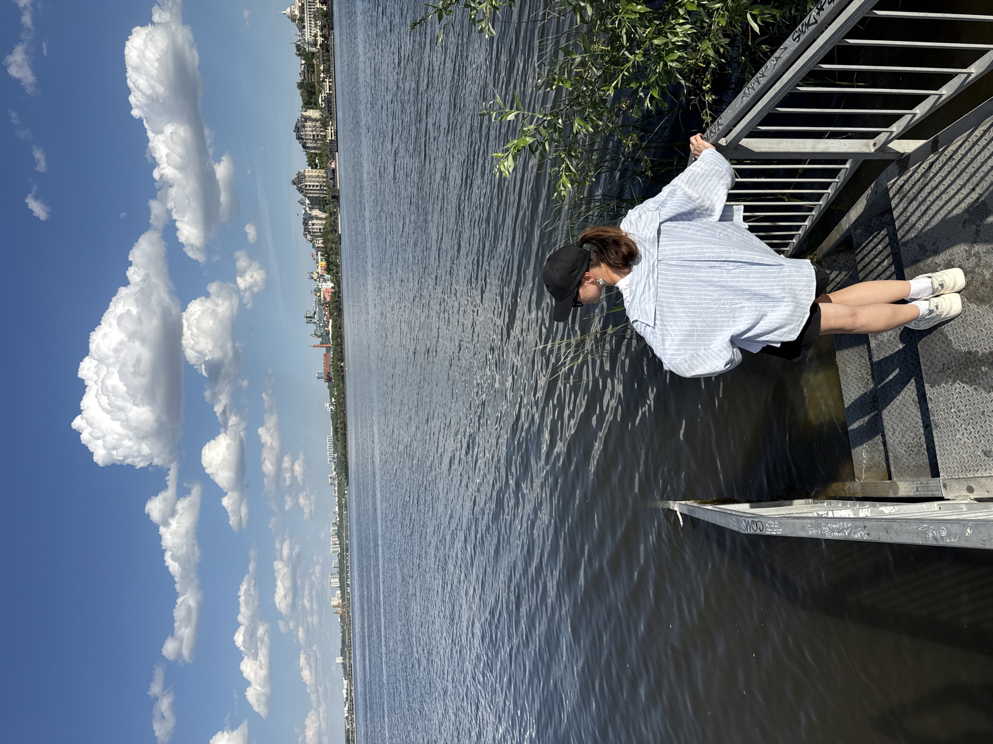 A person is standing on a metal structure extending over water in Kazan, Russia. The person, likely female, is wearing a blue and white striped shirt, dark shorts, white socks, and white shoes. They are also wearing a dark baseball cap and appear to be looking down at the water. The metal structure is a set of steps leading down to the water, with graffiti markings on the lower section that is partially submerged. The water is dark and rippled, reflecting the sky. In the background, across the water, is the Kazan cityscape with various buildings visible. The sky is blue with scattered white clouds. The image is rotated 90 degrees clockwise.