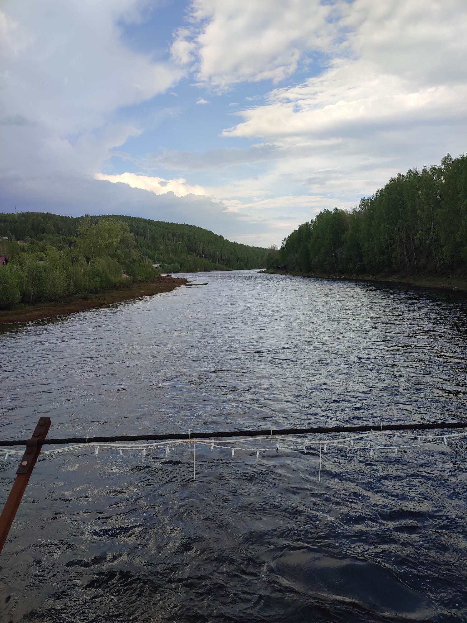 The image depicts a wide river flowing through a forested landscape, likely in the vicinity of Усьва, Russia, though no specific identifying features confirm this location. The river fills most of the frame, and the water is dark and rippled, suggesting a slight current. Both banks are lined with dense green trees and vegetation extending up gentle slopes. The sky is partly cloudy, with patches of blue and gray, indicating a mix of sunny and overcast conditions. In the foreground, a section of what appears to be a cable or rope with hanging white lights or decorations is visible, perhaps part of a bridge or an installation over the water. No people are visible in the scene.