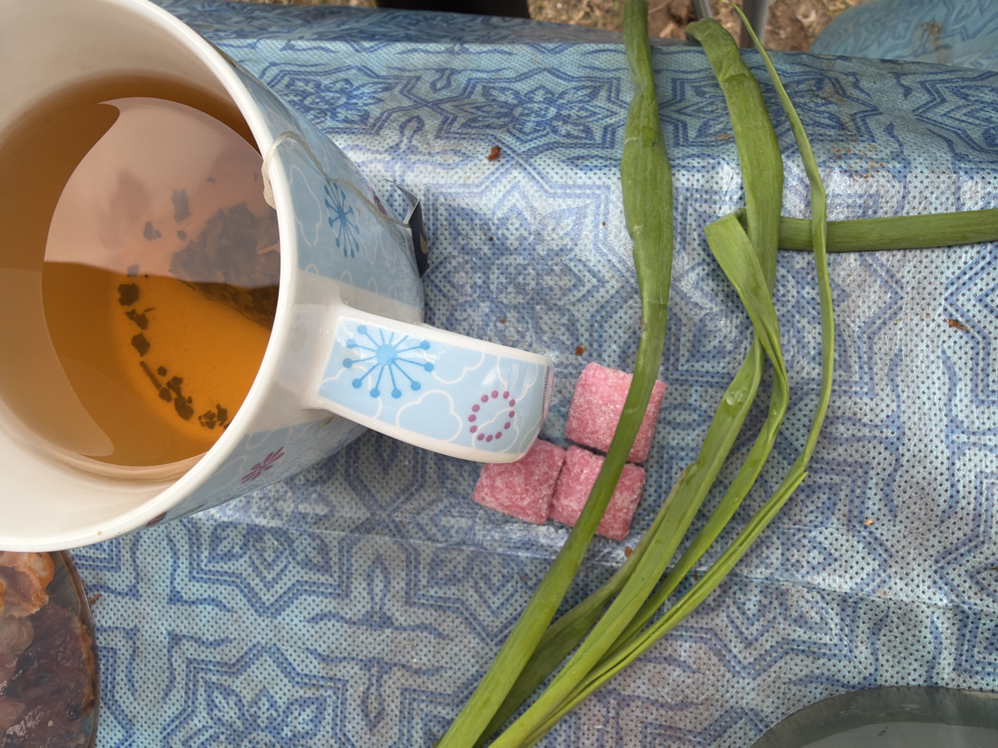 A mug of tea with a tea bag inside sits on a blue and white patterned surface next to three pink sugar cubes and several green onion stalks. The mug has a white interior and a light blue exterior decorated with blue and pink floral patterns. The tea is a light amber color. The pink sugar cubes are positioned near the mug's handle. The green onion stalks are scattered beside the sugar cubes. The surface appears to be a textured tablecloth. The location is Бронница, Russia.