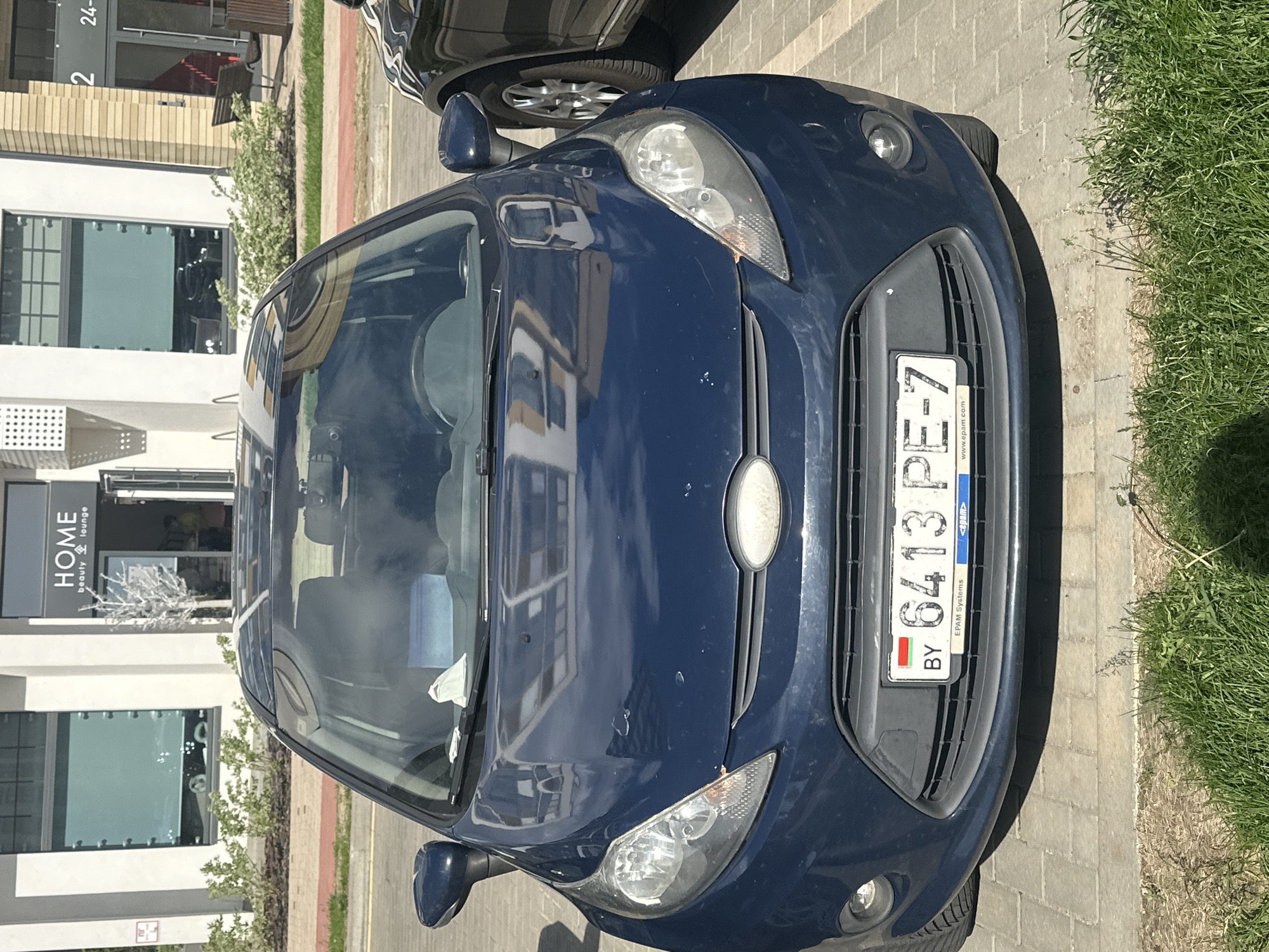 The image depicts the front of a blue car parked on a paved surface. The car's license plate is visible, displaying "6413 PE-7," which indicates it is registered in Belarus. The license plate also mentions "EPAM Systems" and the website "www.epam.com." Behind the car, there is a building with a sign that reads "HOME beauty lounge." To the right of the car, there is a patch of grass. The location is Бараўлянскі сельскі Савет, Belarus.
