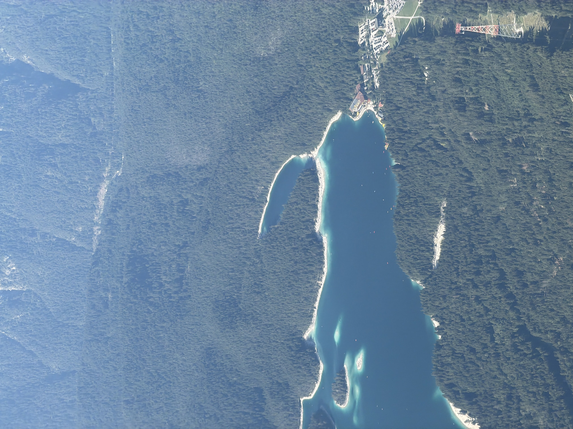 This is an aerial, high-angle view of a picturesque mountain lake surrounded by a dense evergreen forest, likely taken from a high vantage point such as a drone or aircraft. The lake is a vibrant turquoise blue, with a rocky island and several smaller islets dotting its surface. White sand beaches line portions of the lake's edge.
In the mid-ground, a resort complex with multiple buildings and a large parking lot is situated along the lake's shore. The buildings appear to be hotel or lodge-style accommodations. Further into the forest, a red and white lattice tower, possibly part of a ski lift or cable car system, stands tall against the backdrop of the trees. The surrounding mountains are covered in a thick, dark green forest. The overall impression is one of a serene and natural landscape, with human development integrated into the environment. The image is likely taken near Garmisch-Partenkirchen, Germany, a region known for its stunning alpine scenery and recreational opportunities.