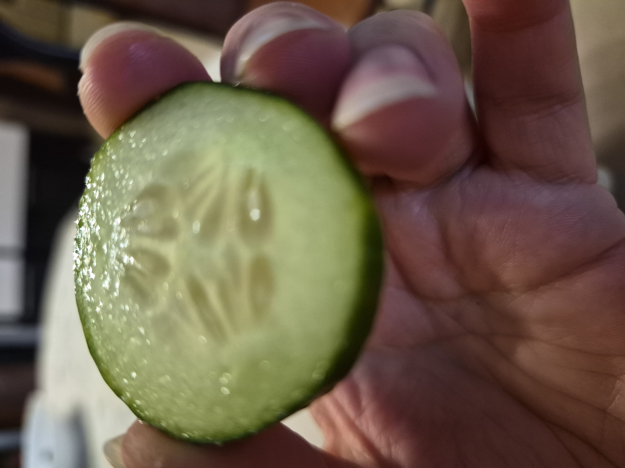 The image shows a close-up of a person's hand holding a slice of cucumber. The cucumber slice is green with visible seeds in the center. The hand is positioned to the right of the cucumber slice, with fingers partially obscuring it. The skin on the hand appears to have lines and texture. The background is out of focus and shows a blurred, warm-toned interior environment, suggesting a domestic setting. There are some bright spots of light reflecting off the cucumber slice, possibly from an artificial light source. The lighting appears to be indoor lighting. No text is visible in the image.