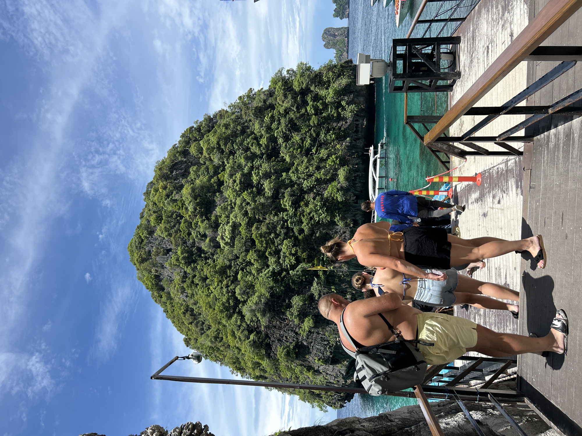The image captures a scene at a pier in Ao Nang, Thailand, under a bright, partly cloudy sky, suggesting a warm, sunny day. The setting is a wooden pier with a railing, overlooking the turquoise waters of the sea. In the background, a lush, green, rocky island rises dramatically from the water, a common sight in the limestone karst landscape of the region. Several boats are visible docked at the pier or in the water, hinting at a tourist or transportation hub.
The foreground features several people, seemingly tourists, dressed in beachwear. One person in the front is wearing light yellow shorts and a backpack, facing away from the camera. Behind them, two individuals, a woman in a bikini and denim shorts and another woman in a bikini and black shorts, are also partially visible, facing the same direction. Further back, a person in a blue shirt with the word "SERVICE" clearly printed on the back is standing, possibly a staff member. The overall atmosphere appears relaxed and vacation-like, with people possibly waiting to board a boat or having just disembarked. The clear water and vibrant greenery of the island contribute to a tropical, scenic ambiance.