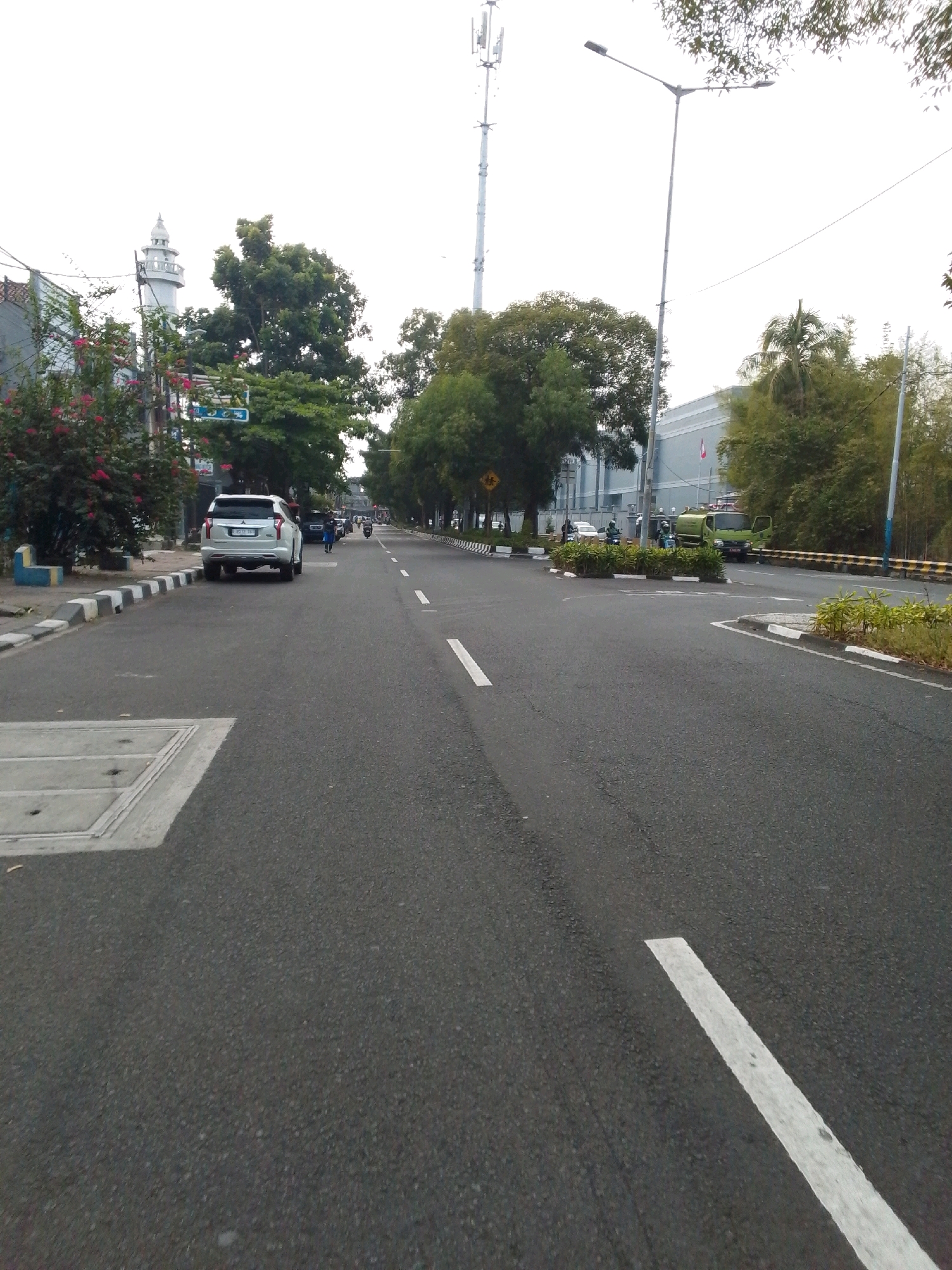 The image shows a wide, paved road in East Jakarta, Indonesia, with a cloudy sky overhead. On the left side of the road, a white SUV is parked at the curb, which is lined with black and white checkered concrete blocks. Adjacent to the parking area is a small garden with red flowers. Further down the street, a white minaret of a mosque is visible.
The road itself is marked with white dashed lines and a solid white line on the right side. A street light pole with two lamps stands tall on the right side of the road, accompanied by a taller telecommunications tower in the distance. In the middle ground, a green truck with its door open is parked, and several people on motorcycles are present. Lush green trees line both sides of the road, with a modern building visible behind some of the trees on the right. The overall impression is a typical urban street scene in Indonesia.