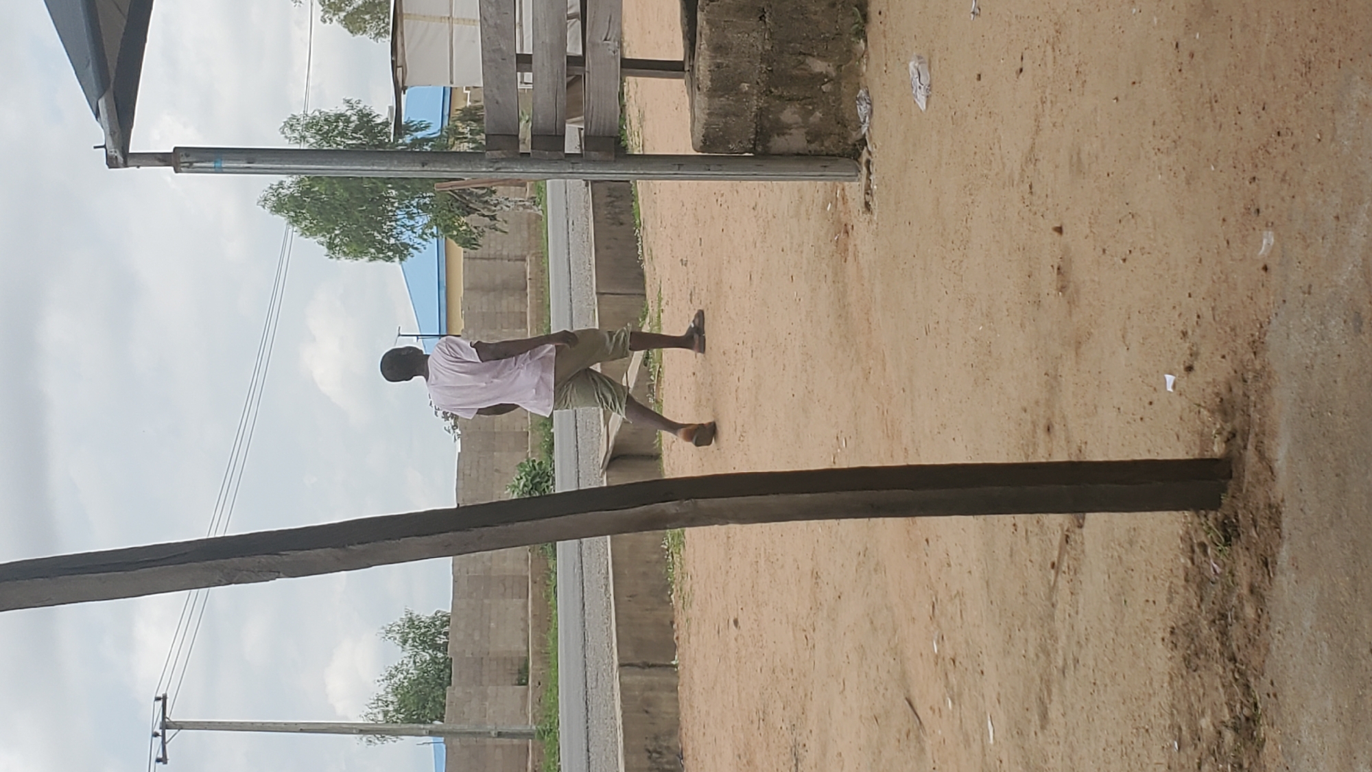 A person walks away from the camera in an outdoor setting, likely in Jalingo, Nigeria, under a cloudy sky. The individual, a dark-skinned male, wears a light pink t-shirt and olive-green shorts. He is walking on a sandy or dirt surface, with a concrete curb and road visible in the background. To the right of the frame, a wooden structure with a metal support pole is present. A utility pole with power lines is visible on the left side of the image. The general environment appears somewhat undeveloped, with some scattered debris on the ground.