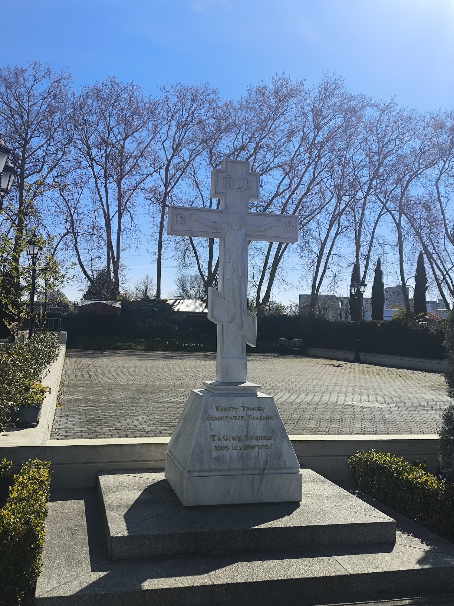 The image shows a white Orthodox cross with the inscription "Кресту Твоему поклоняемся, Владыко! Ta Groix, Seigneur, nous la vénérons!" located in Strasbourg, France. The cross stands on a multi-tiered stone base in a paved area with trees in the background. Lamp posts line the perimeter. The sky is clear and blue.