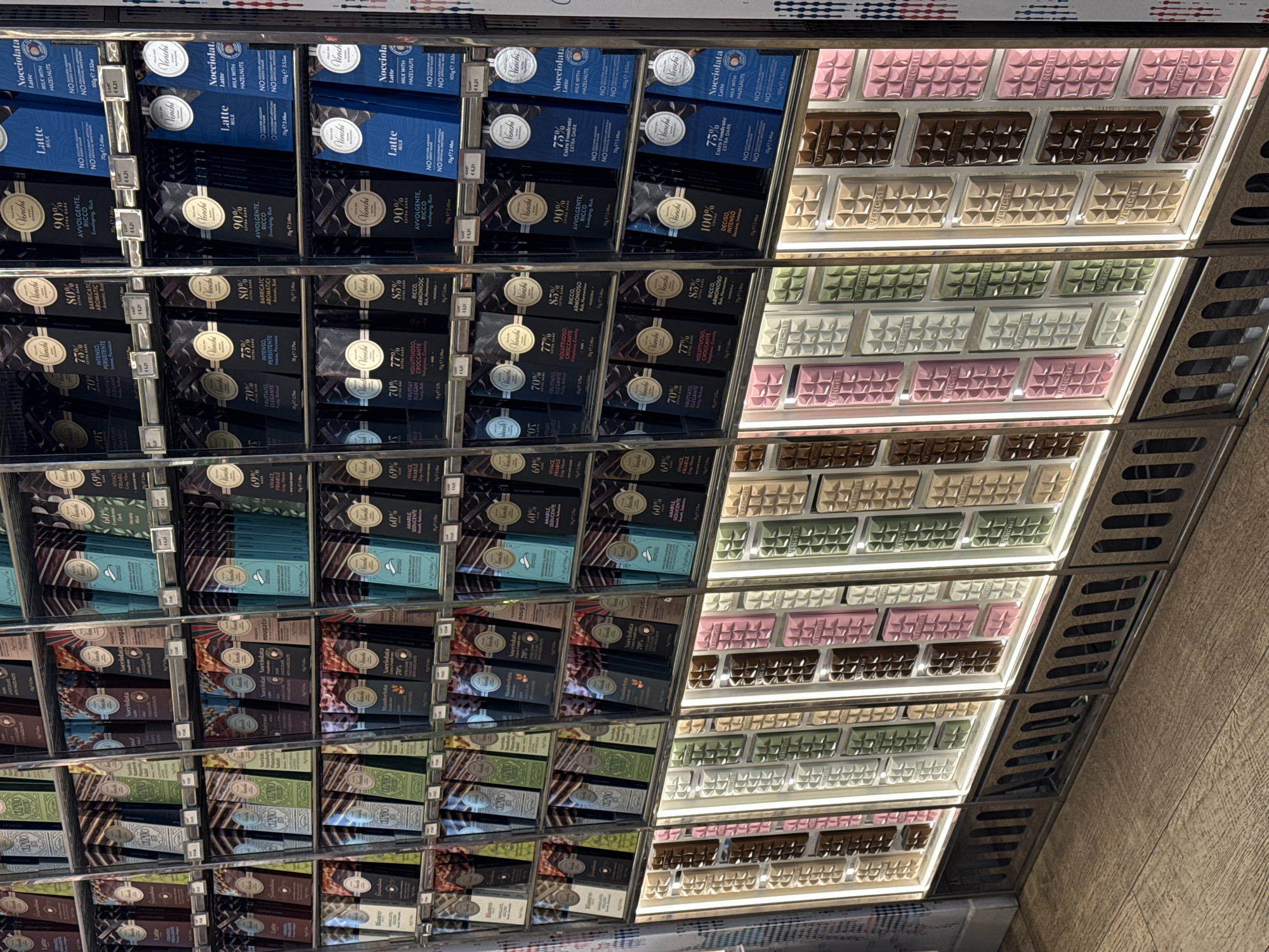 This photograph, taken indoors at an oblique angle, showcases a well-organized display of Venchi chocolate products in Noventa di Piave, Italy. The products are arranged on multiple shelves in a retail environment. On the left, the shelves are lined with packaged chocolate bars of varying percentages (60% to 100%) and flavors, including 'Latte', 'Nocciolata', and 'Bianco'. On the right, there are rows of unwrapped Venchi chocolate bars in pink, brown, white, and green hues. Price tags accompany each product, and the shelf structure is metal, complete with cooling vents. The lighting is bright and artificial. The setting is a typical retail space for chocolates.