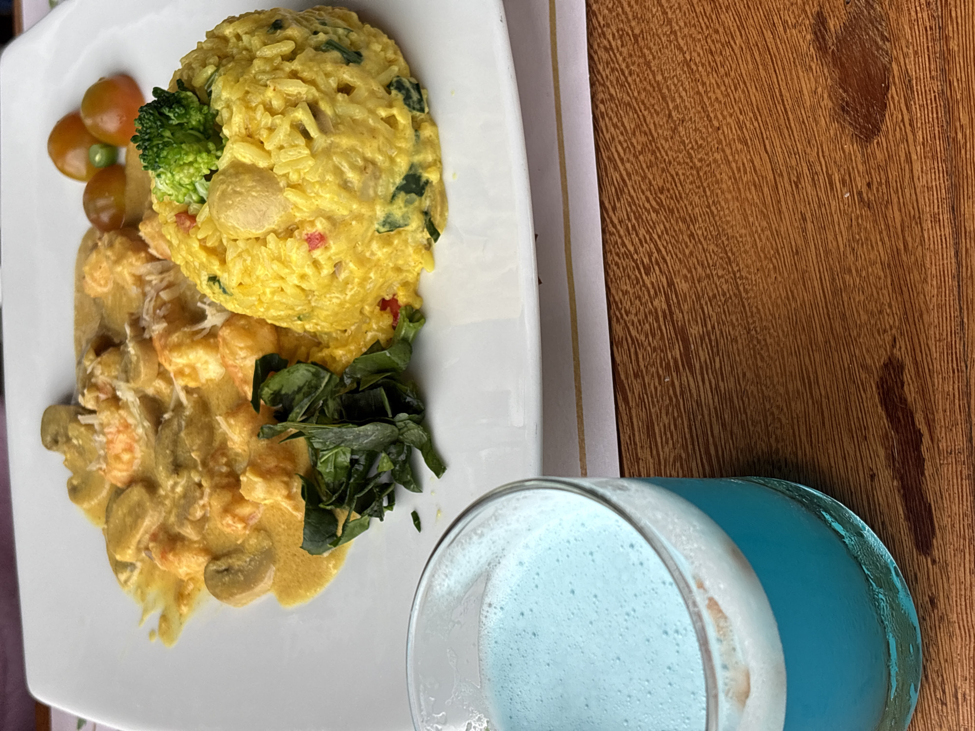 The image shows a plate of food on a white surface, next to a glass of blue liquid. The food consists of a creamy shrimp and mushroom dish, a mound of yellow rice with greens and broccoli, and a few cherry tomatoes. The blue liquid in the glass appears to be a beverage, possibly a smoothie or cocktail. The setting is likely a restaurant, given the presentation of the food and drink. The image is taken from a slightly elevated angle, looking down at the plate and glass. The background is blurred, but suggests a wooden table.