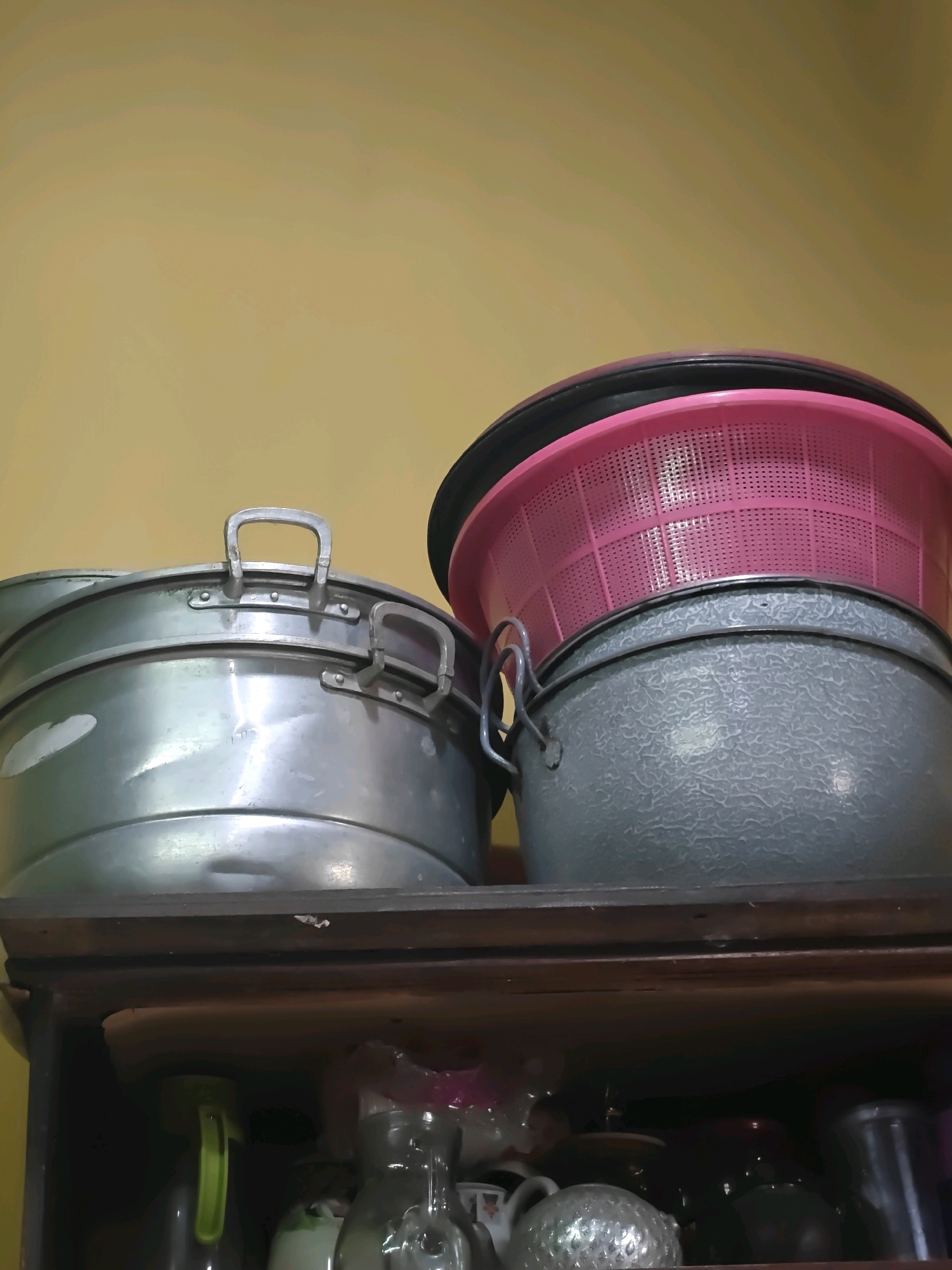 A close-up, low-angle shot captures two stacked kitchen basins on a wooden shelf against a yellow wall. The top basin is a large, silver-toned metal pot with two handles. Beneath it rests a smaller, gray, textured basin. Perched on top of the gray basin is a pink plastic colander with a black rim. The shelf below the basins is visible, displaying a dark interior with various kitchen items, including a green spray bottle, a glass bottle, and other containers. The scene appears to be in a kitchen in Sumberwono, Indonesia.