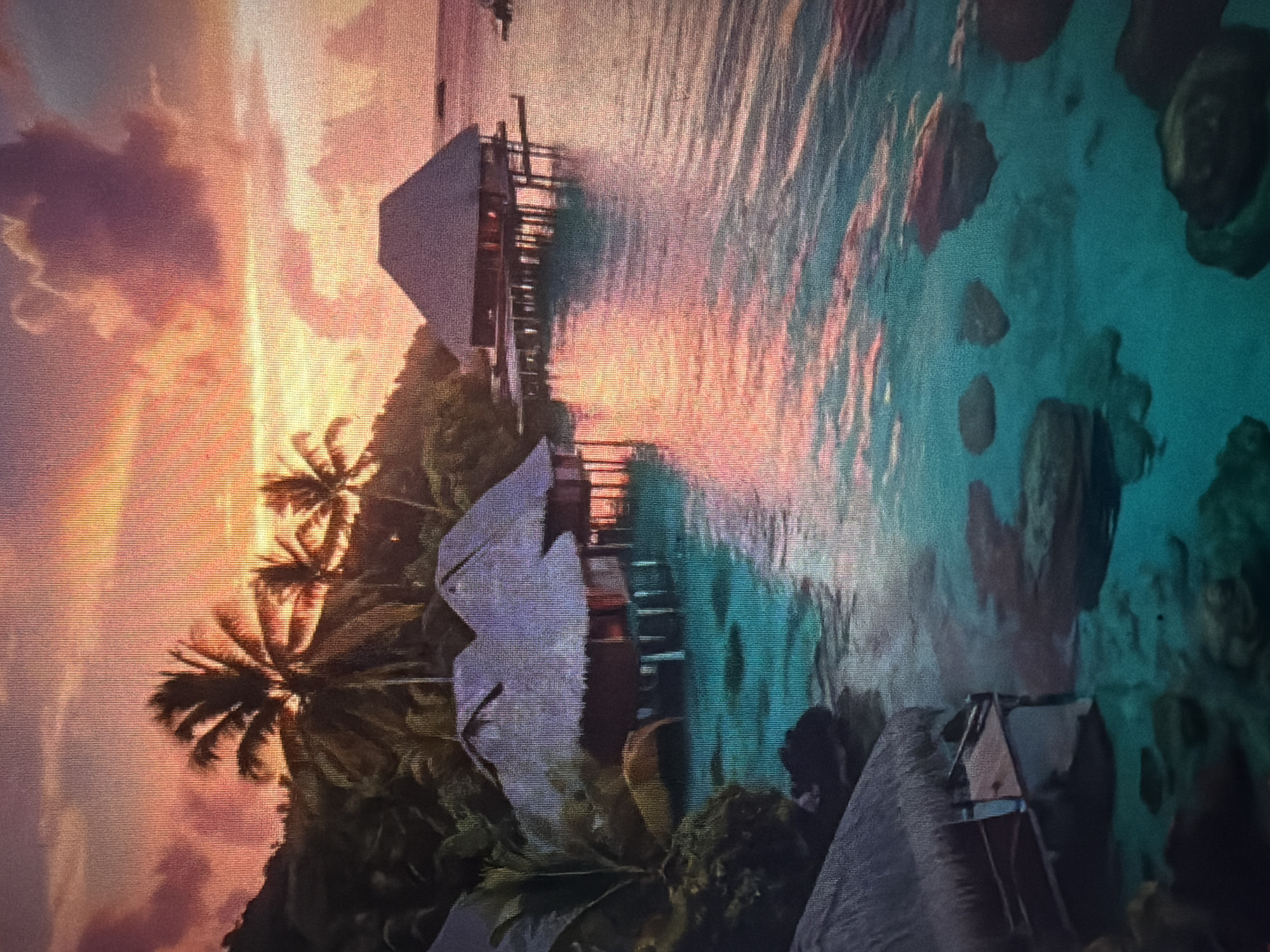 This image depicts an overwater bungalow resort at sunset. The sky is ablaze with hues of orange, pink, and purple, with dramatic clouds reflecting on the calm, turquoise water. Several thatched-roof bungalows are visible, built on stilts over the shallow, clear ocean. The bungalows have dark wooden exteriors and appear to be lit from within, casting a warm glow. Palm trees and lush green foliage flank the shoreline in the background. Rocks are visible submerged in the water near the bungalows. The scene conveys a sense of tropical tranquility and luxury. The location is identified as Baan Mae Nam, Thailand. There are no people visible in the image.