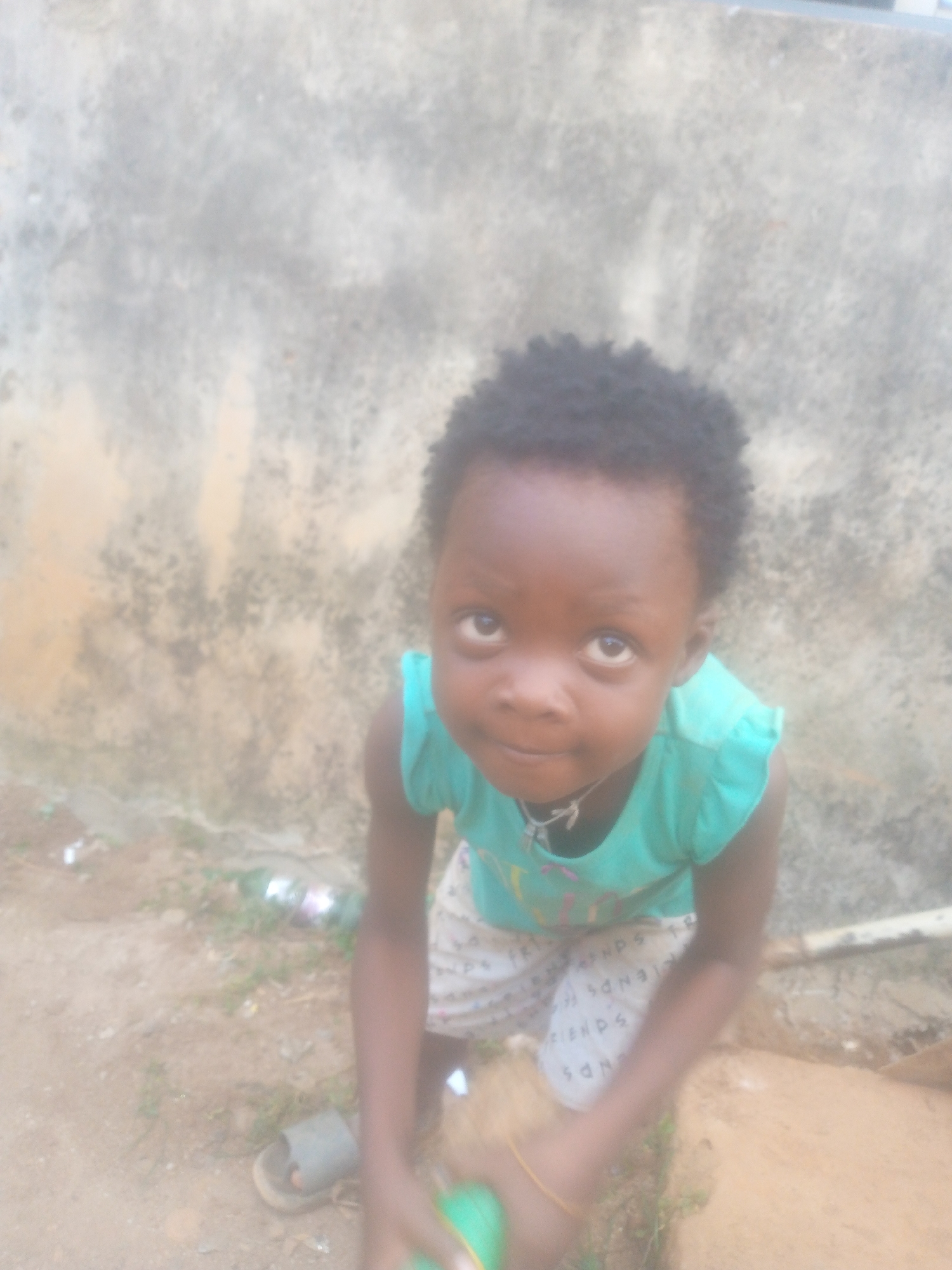 A young child with dark skin and short, curly hair is looking up at the camera. The child is wearing a turquoise top and patterned shorts. The background consists of a textured wall and a dirt ground with some debris. One sandal is visible on the child's left foot. The location is Unknown, Unknown.