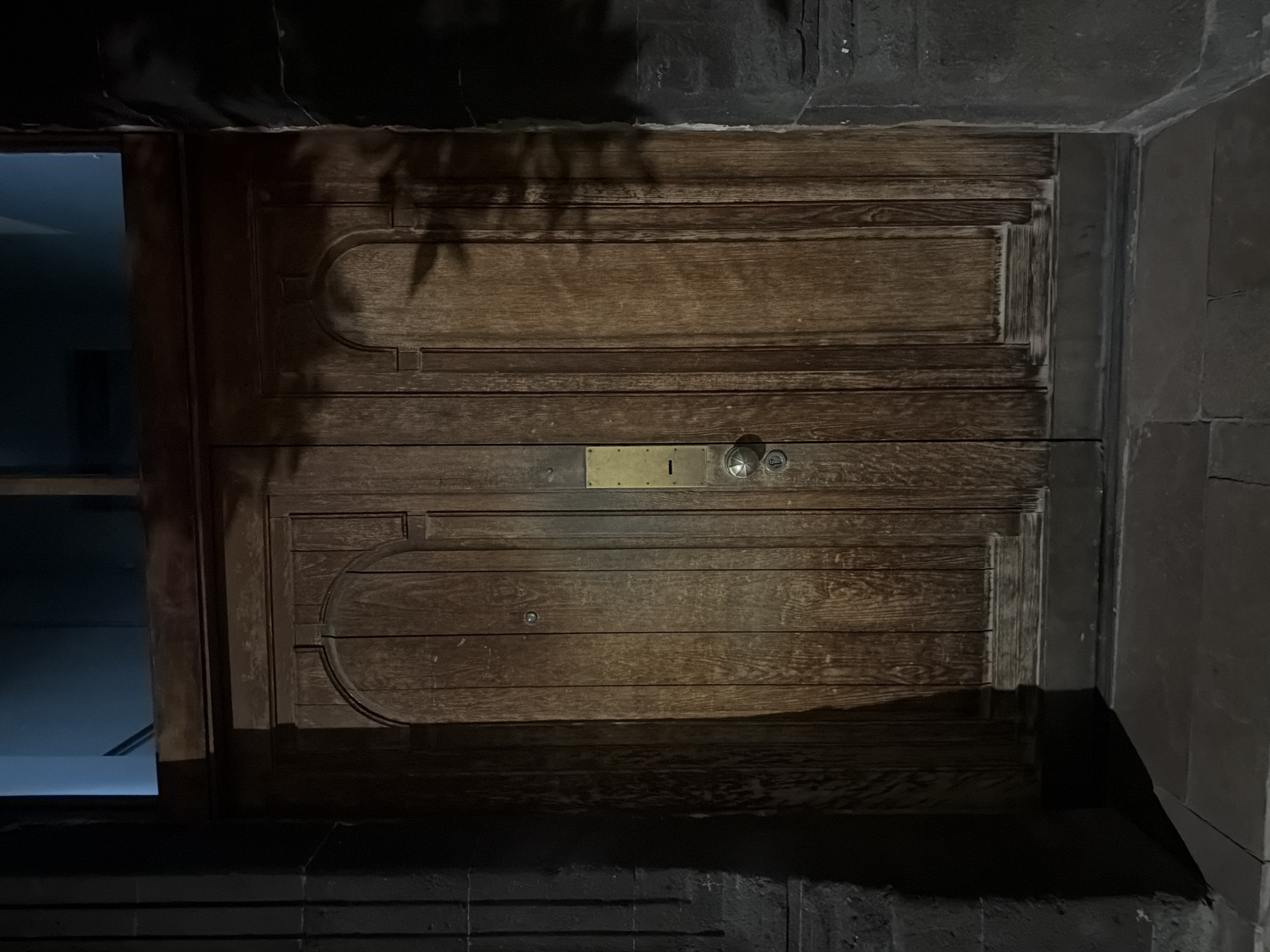 The image shows a dark, wooden, double door set into a stone wall. The door has two horizontal sections with rounded decorations on the top and bottom of each. There is a brass plate with a keyhole and a knob on the center of the door. The door appears to be located at night, possibly an entrance to a building. The location is in Yerevan, Armenia.