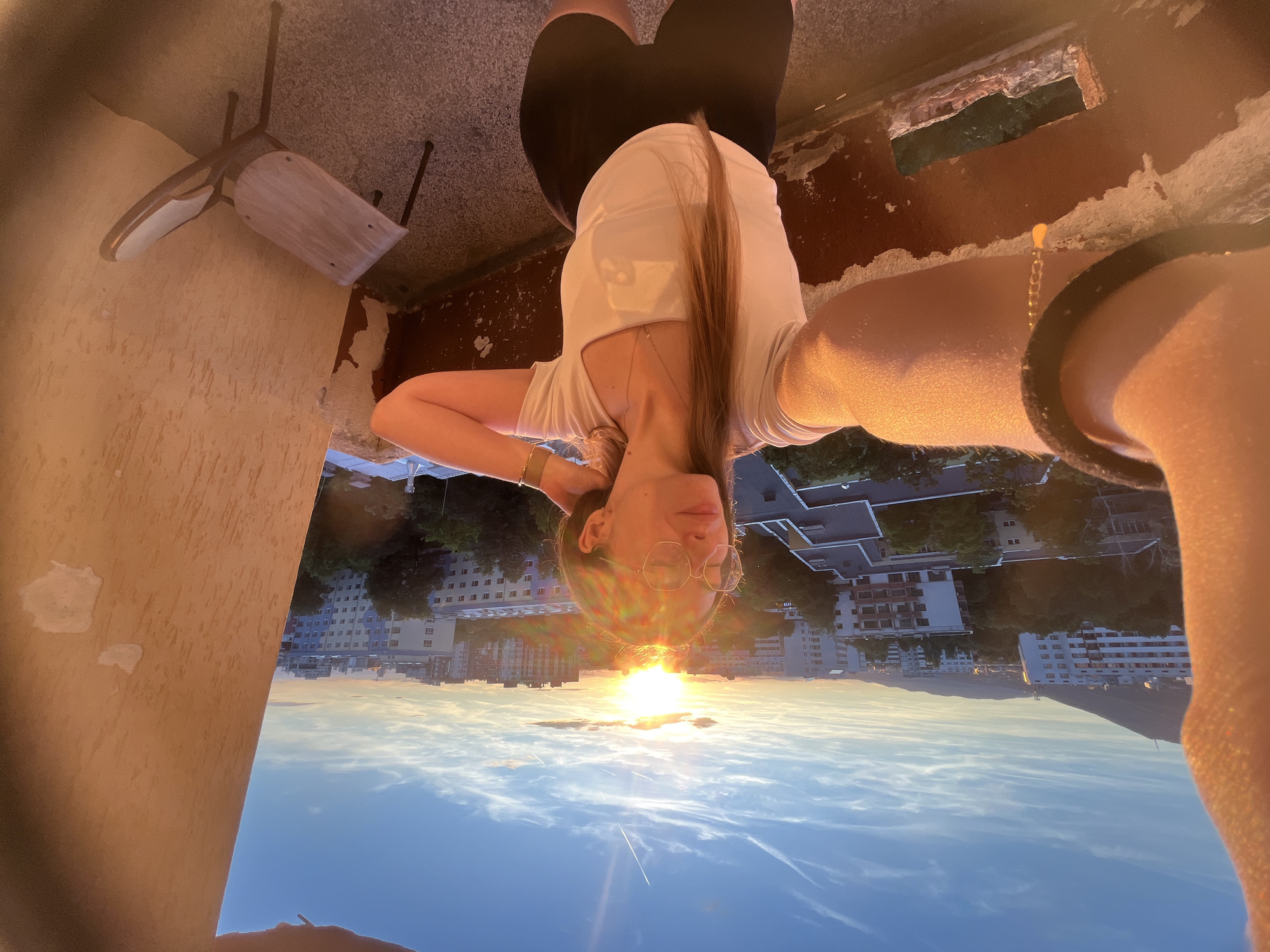 In the city of Unknown, Unknown, a young woman with long blonde hair and glasses stands on a balcony, taking a selfie at sunset. The sun is low in the sky, casting a warm golden light over the scene and creating a lens flare. The woman is wearing a white t-shirt and black shorts. She is touching her hair with her right hand and has her left arm extended to take the photo. In the background, a cityscape is visible, with apartment buildings and trees under a blue sky with wispy clouds. A single chair sits on the balcony to the woman's right. The balcony railing is visible in the foreground, showing signs of wear and tear.
