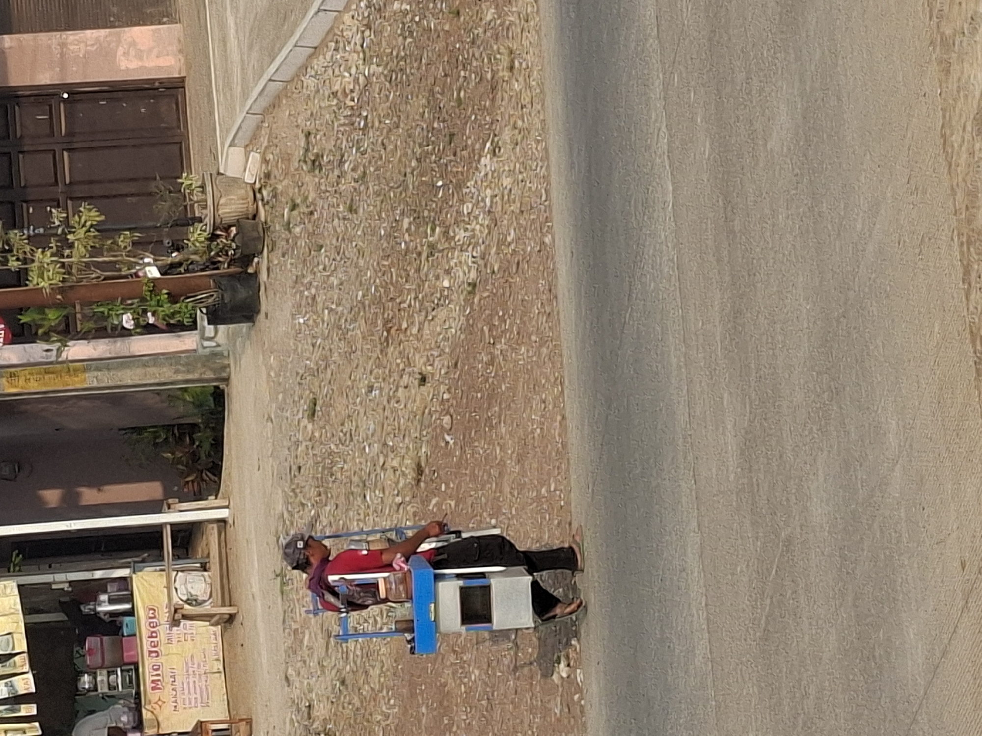 A person wearing a red shirt and a cap walks along a road in Ciheulang Tonggoh, Indonesia. They are carrying a mobile cart with a blue frame and a grey rectangular body, which appears to be a food or beverage cart. The cart has several compartments and containers. The person is walking on the right side of a paved road, with a gravelly area on their left, which leads to a building with a yellow awning. The building has a sign that reads "Mio Jebo". The environment is outdoors with natural lighting, suggesting it is daytime. The scene is captured from a side profile, showing the person and their cart in motion.