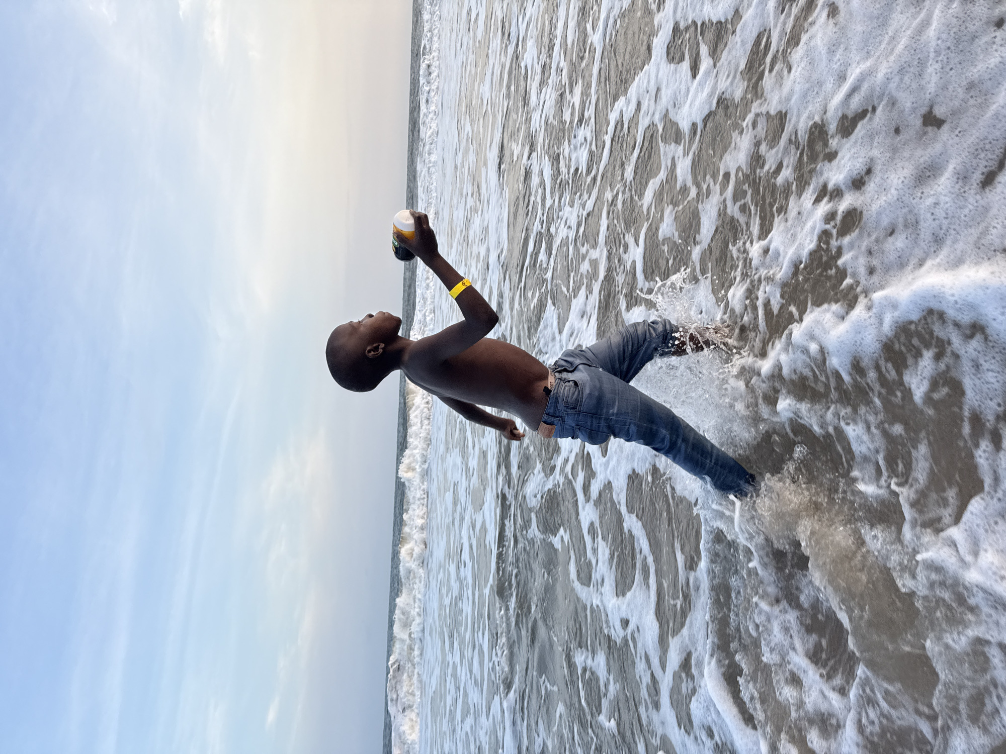 A young, dark-skinned person wearing blue jeans stands in the surf on a beach, possibly near Mopo Onijebu, Nigeria. The person is holding a container to their lips and appears to be drinking. They are wearing a yellow wristband. The sea foam churns around their legs. The sky is visible in the upper-left corner of the image.