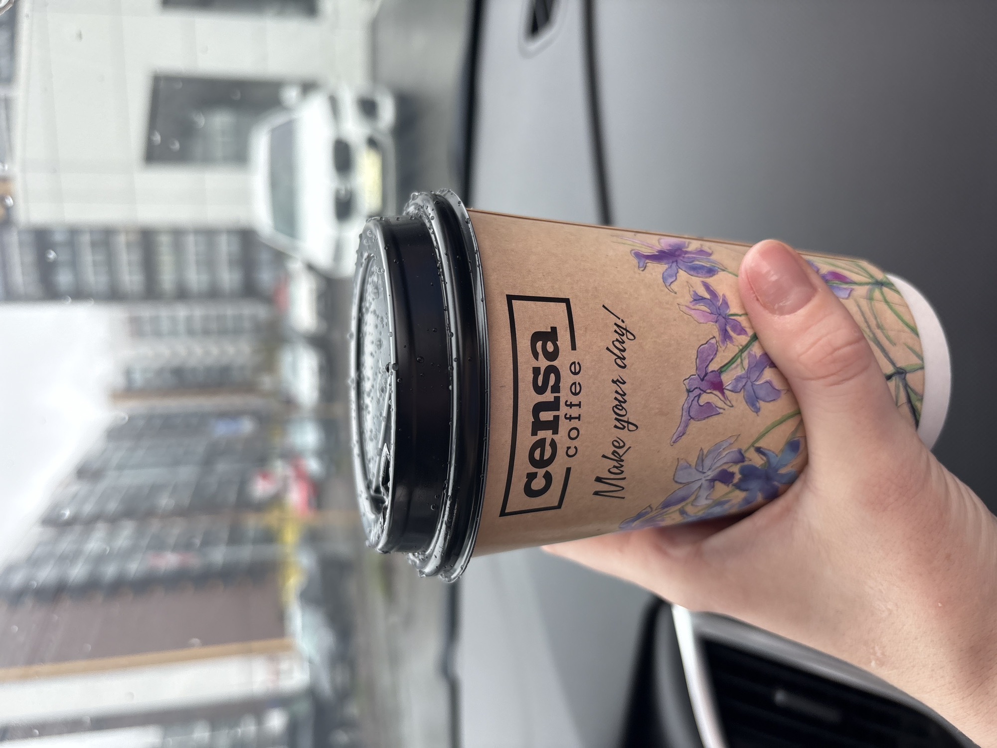 A person's hand holds a brown coffee cup with a black lid inside what appears to be a vehicle. The cup is adorned with a purple floral design and features the text "censa coffee" in a black outlined box and "Make your day!" in a handwritten-style font below it. Numerous small water droplets are visible on the black lid and the outer surface of the cup, suggesting rain or condensation.
The background, viewed through a window, is blurred, showing an urban environment with buildings and possibly other vehicles, consistent with a street or parking area. The presence of water droplets on the window surface further indicates rainy or wet weather conditions outside. The overall lighting suggests it is daytime, likely overcast. Elements of a car's interior, such as a dashboard or door panel, are visible on the right side of the frame, confirming the setting inside a vehicle. The scene takes place in Kazan, Russia.