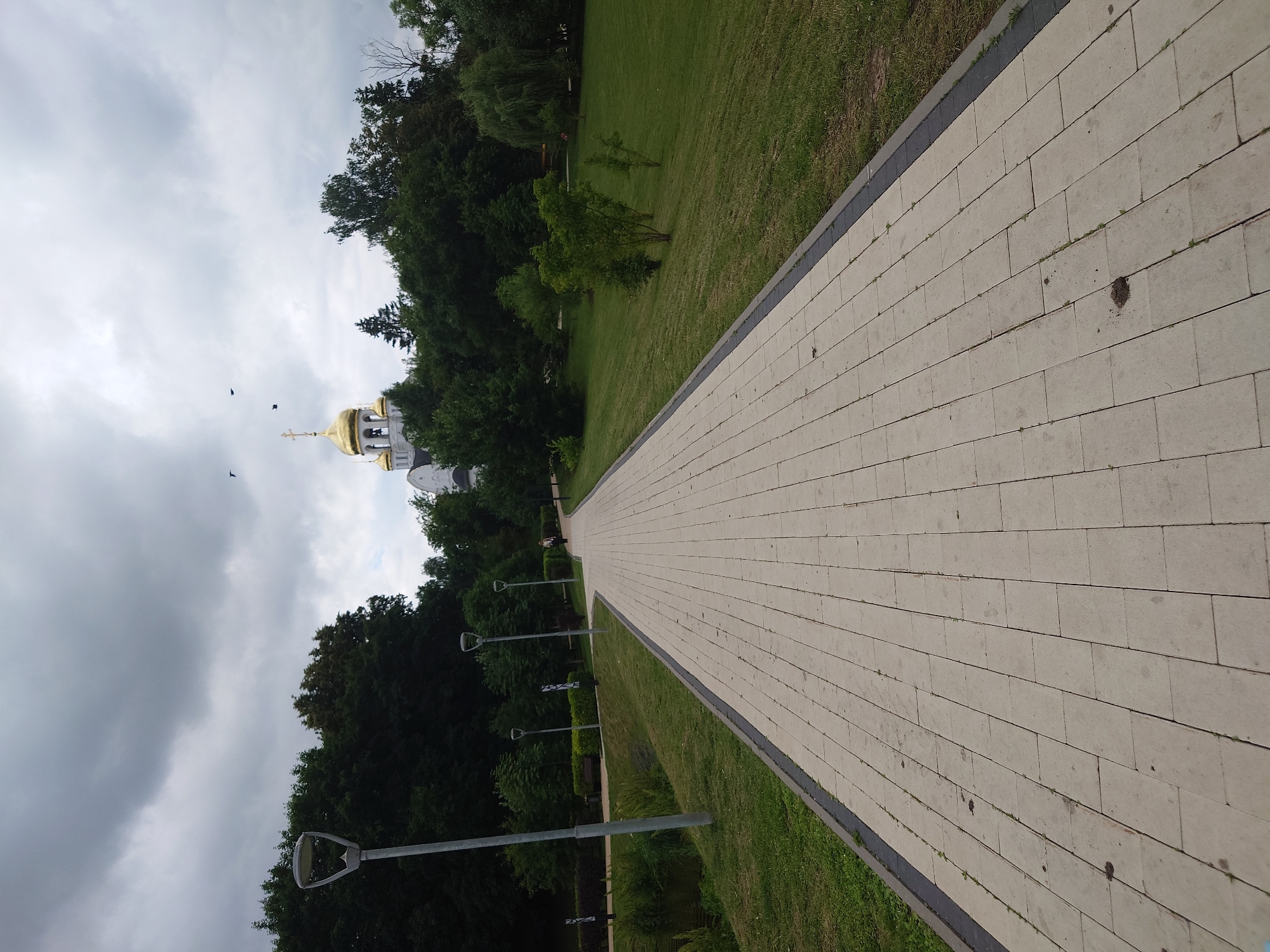 A person is walking on a paved pathway in a park on a cloudy day. The pathway is made of light-colored rectangular stones and is bordered by a dark strip. To the left of the pathway is a grassy area with a small body of water. To the right of the pathway is a sloping lawn with trees. In the background, there is a white building with golden domes, identified as the Church of the Resurrection of Christ in Kommunarka, Russia. Several streetlights are visible along the pathway. There are also a few birds in the sky.