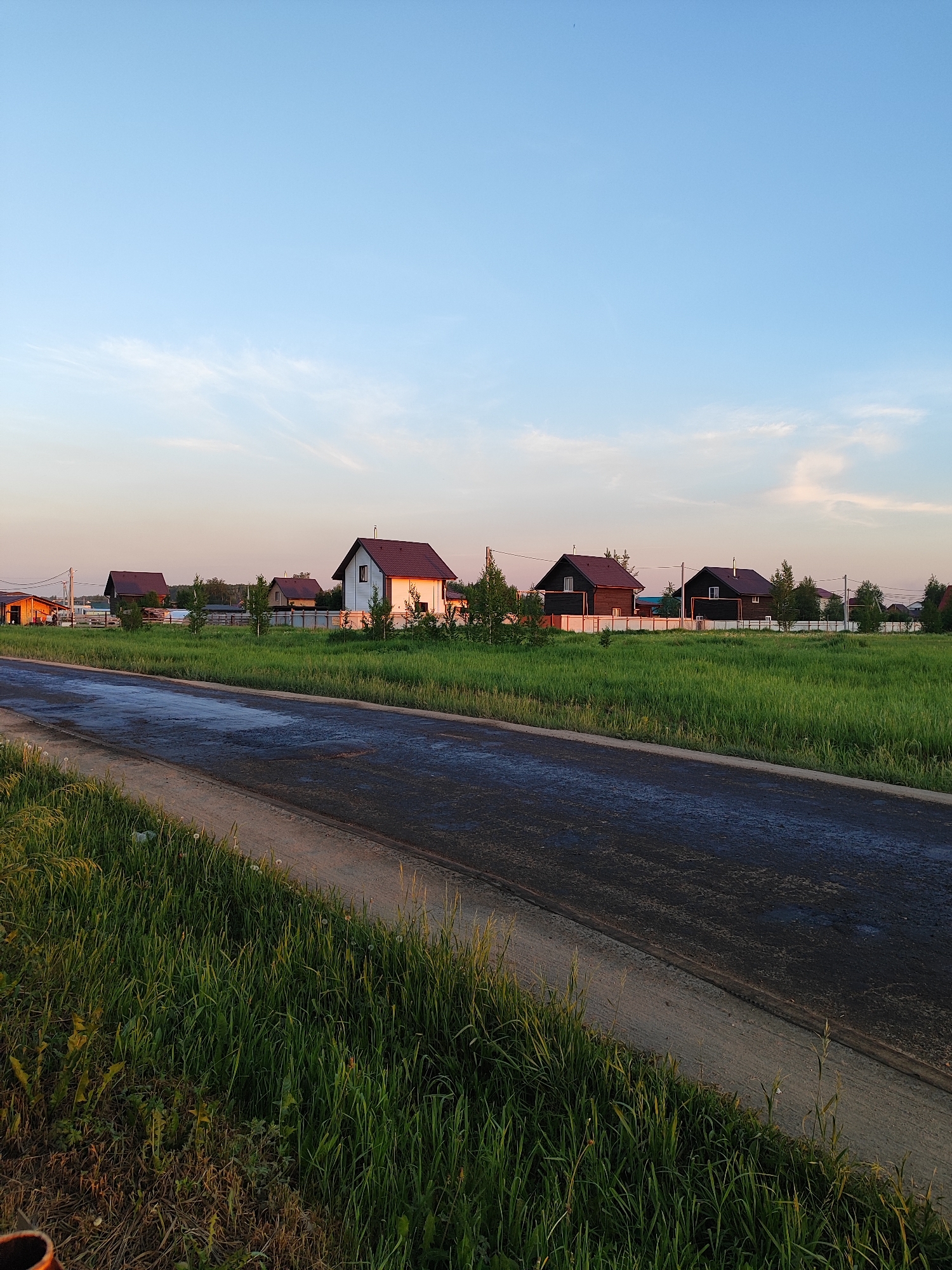 The image shows a rural landscape with several houses situated behind a grassy field. A paved road is in the foreground, with grass growing along its edge. The sky is clear with some wispy clouds, suggesting it might be dusk or dawn. The houses appear to be modern and of similar design, with dark roofs and light-colored walls. The environment is serene and undeveloped. The location is in the city of Marusino, Russia.