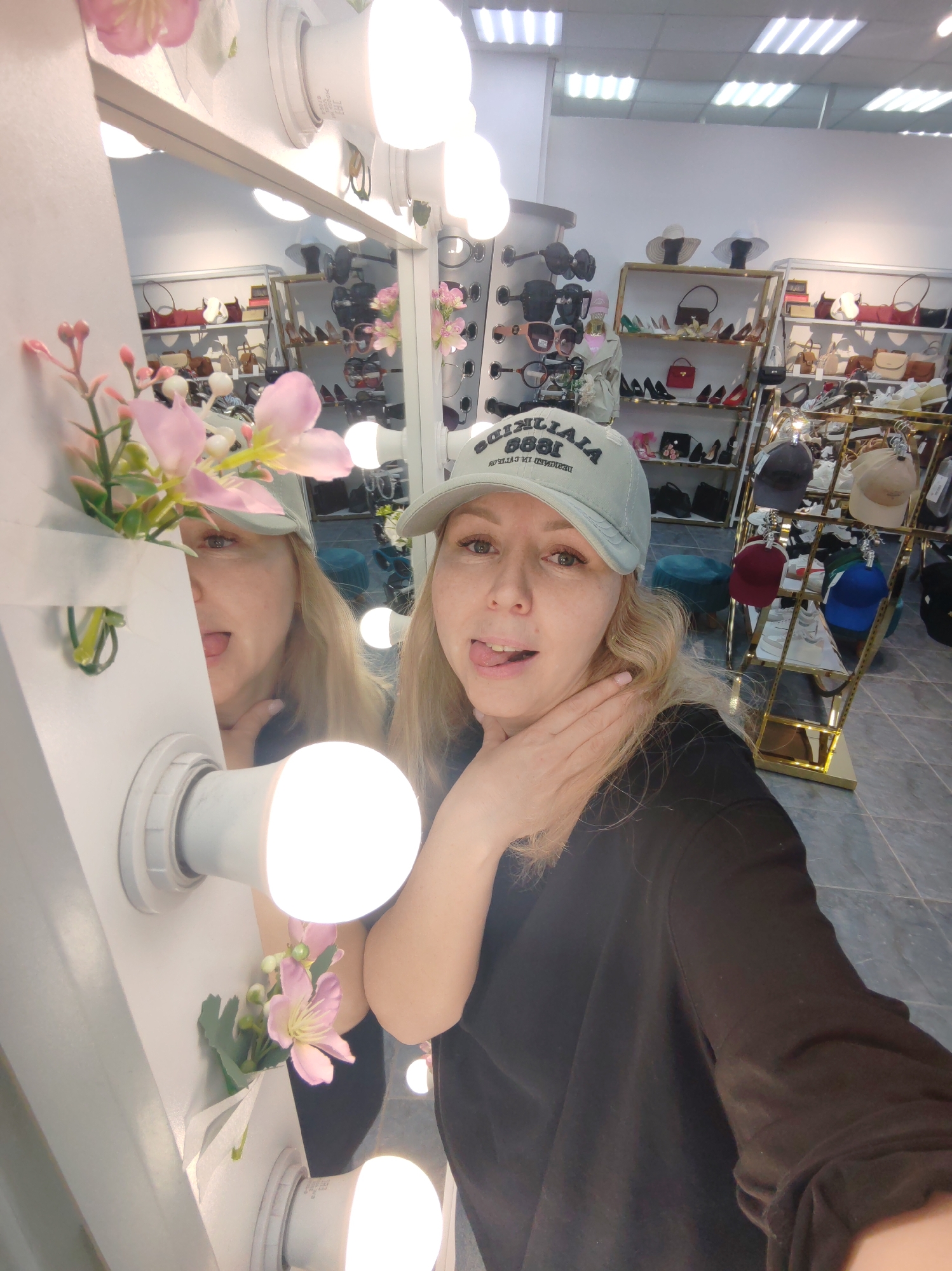 The image is a selfie taken in what appears to be a retail store. A woman wearing a light blue cap with "QUILILLA 1888" inscribed on it is the primary subject. She is sticking out her tongue and has her hand near her neck. Part of her reflection is visible in a mirror with bulb lights and artificial flowers. The background shows racks filled with shoes, bags, and hats. Based on the request, the location is in Chaikovsky, Russia, but this cannot be verified from the image alone.