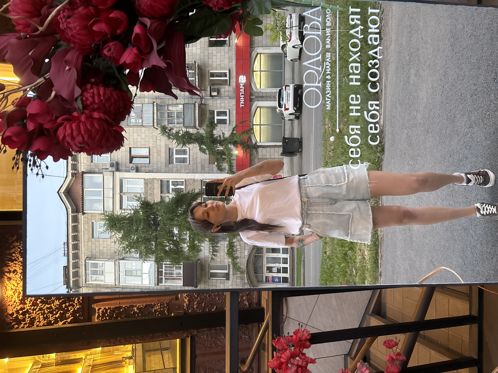 A young woman with her hair in a ponytail stands in front of a full-length mirror, taking a selfie. She is wearing a white t-shirt and light-wash denim shorts. Her arms are casually resting at her sides, with her right hand in her pocket. She has tattoos on her right arm. The mirror reflects a street scene in Novokuznetsk, Russia, featuring a multi-story apartment building, a tree, two white cars, and a black trash bin. There is a red sign above two arched storefronts that reads "TNH1AM". Below this, in white lettering on the mirror, is the name "ORAOBA" followed by "МАГАЗИН & НАРАЩ. ВАЖНЕЙШЕ ВОЛО". Further down, also in white lettering, are the phrases "себя не находят" and "себя создают". To the left of the mirror, a glimpse of red flowers and a staircase can be seen.