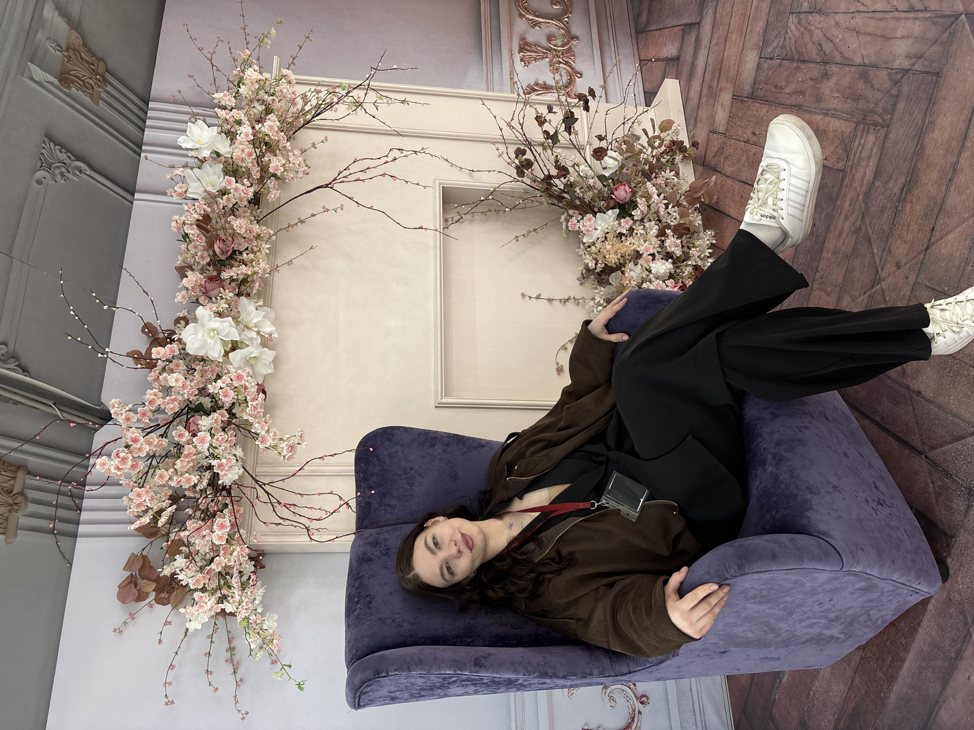 The image displays a young woman lying comfortably on a purple upholstered armchair in a richly decorated indoor setting. She has dark hair and a pleasant, relaxed expression, looking towards the viewer. She is dressed in a dark brown jacket, a black top with a red lanyard visible, and black wide-leg trousers. Her feet, clad in white sneakers, are visible, with the brand "Adidas" discernible on the side of one shoe. Her left hand rests casually on the armrest of the chair.
The background features an elaborate floral arrangement, primarily composed of delicate light pink and white blossoms, resembling cherry blossoms, intertwined with bare branches and some darker, dry foliage. This floral display frames a cream-colored decorative architectural feature, possibly a non-functional fireplace opening or an archway, adorned with classic moldings. The surrounding walls are further embellished with intricate white and ornate golden-bronze decorative moldings and pilasters, suggesting an elegant, classical interior. The floor is covered with dark wooden parquet flooring, laid in a herringbone pattern. The scene depicts a posed shot within a studio or a lavishly decorated room, likely in Perm, Russia. No specific time of day or weather is evident.