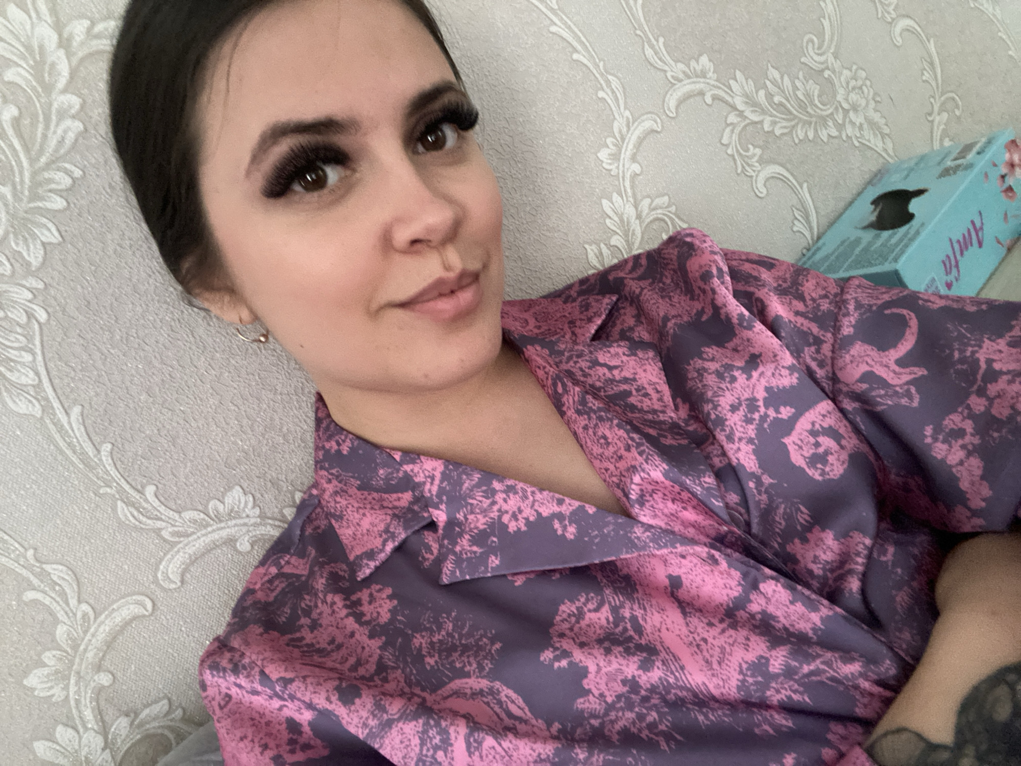 An indoor, close-up photograph captures a woman in a relaxed pose, possibly taking a selfie. She has dark hair pulled back, prominent dark eyelashes, brown eyes, and fair skin, with a small hoop earring visible on her left ear. She wears a pleasant, slightly smiling expression while looking directly at the camera. Her attire is a collared shirt or robe, made of a silky fabric, patterned with an intricate, abstract design in shades of purple and pink.
The background features light grey or beige textured wallpaper adorned with a raised, ornate white floral and swirling pattern. In the upper right corner, a light blue box is visible, tilted against the wall. The word "Amfa" is written vertically in white text on its side, and a dark, stylized silhouette, possibly a bird or abstract symbol, is depicted on its front face. The overall setting appears to be a comfortable indoor space, likely a home. No specific visual cues within the image confirm the provided location context of Chaikovsky, Russia.