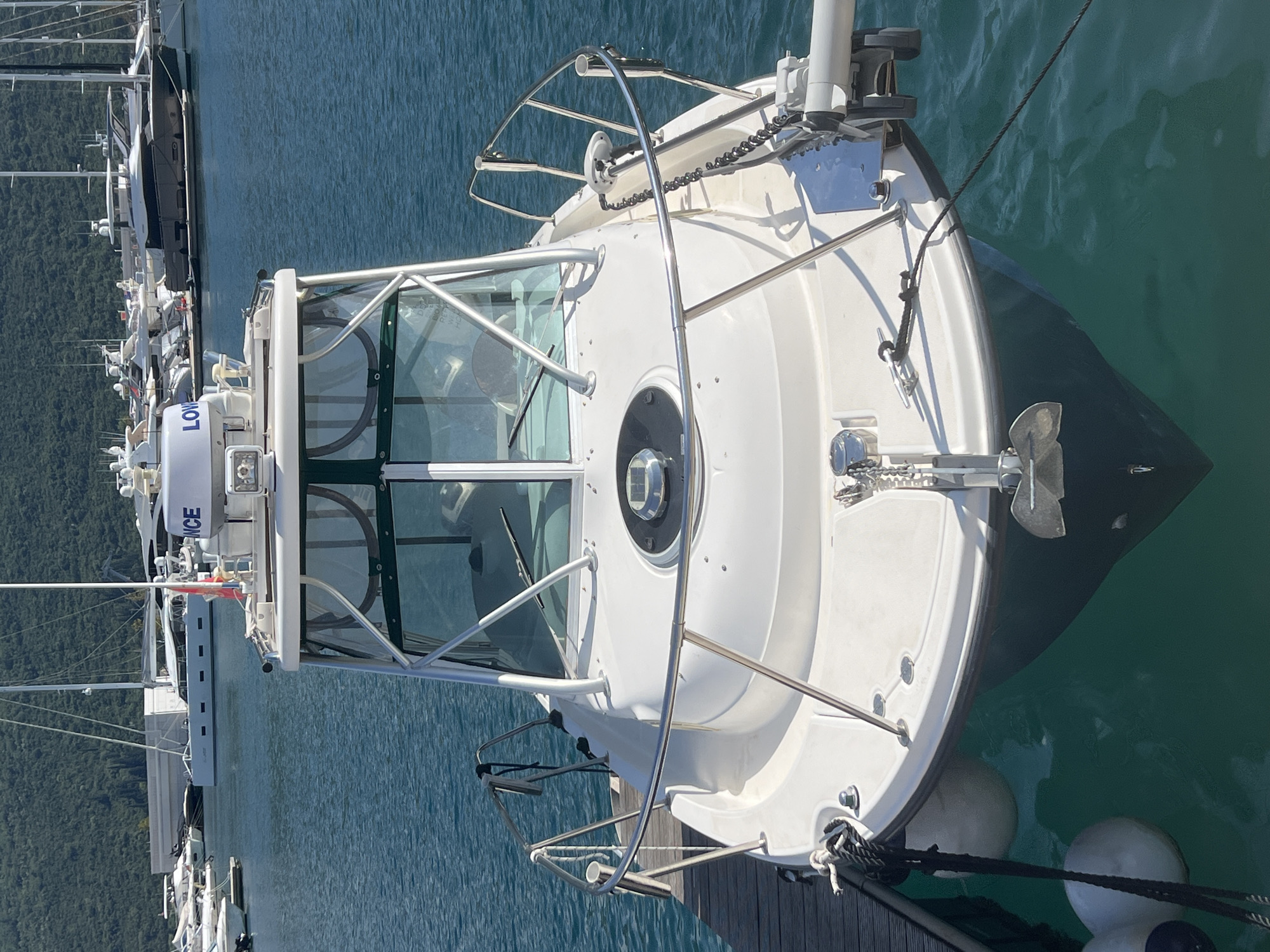 An elevated perspective captures the bow and forward deck of a white powerboat docked in a marina. The vessel, likely a sportfisher or cruiser, is clean and well-maintained, featuring a white hull with a dark blue or black antifouling paint visible below the waterline in the clear, greenish-blue water.
At the very front, a shiny anchor is secured to the bow, with its chain leading to an anchor windlass. Stainless steel bow rails curve along the foredeck. The boat's cabin or pilothouse features a prominent, angled windshield equipped with two wipers. More stainless steel grab rails are mounted on and around the cabin structure. Perched atop the cabin roof is a white radar dome with the brand name "LOWRANCE" clearly legible in blue lettering. Other electronic equipment and masts are also visible on the roof.
The boat is moored to a wooden dock, with black mooring lines and white fenders visible, securing it in place. In the background, to the left of the main vessel, other boats are docked in the tranquil marina. Beyond the water, lush, green, tree-covered mountains rise steeply, suggesting a picturesque coastal or fjord-like setting. The scene is bathed in bright daylight, indicating clear and sunny weather, with calm water reflecting the blue sky. The image conveys a peaceful atmosphere in a scenic location.