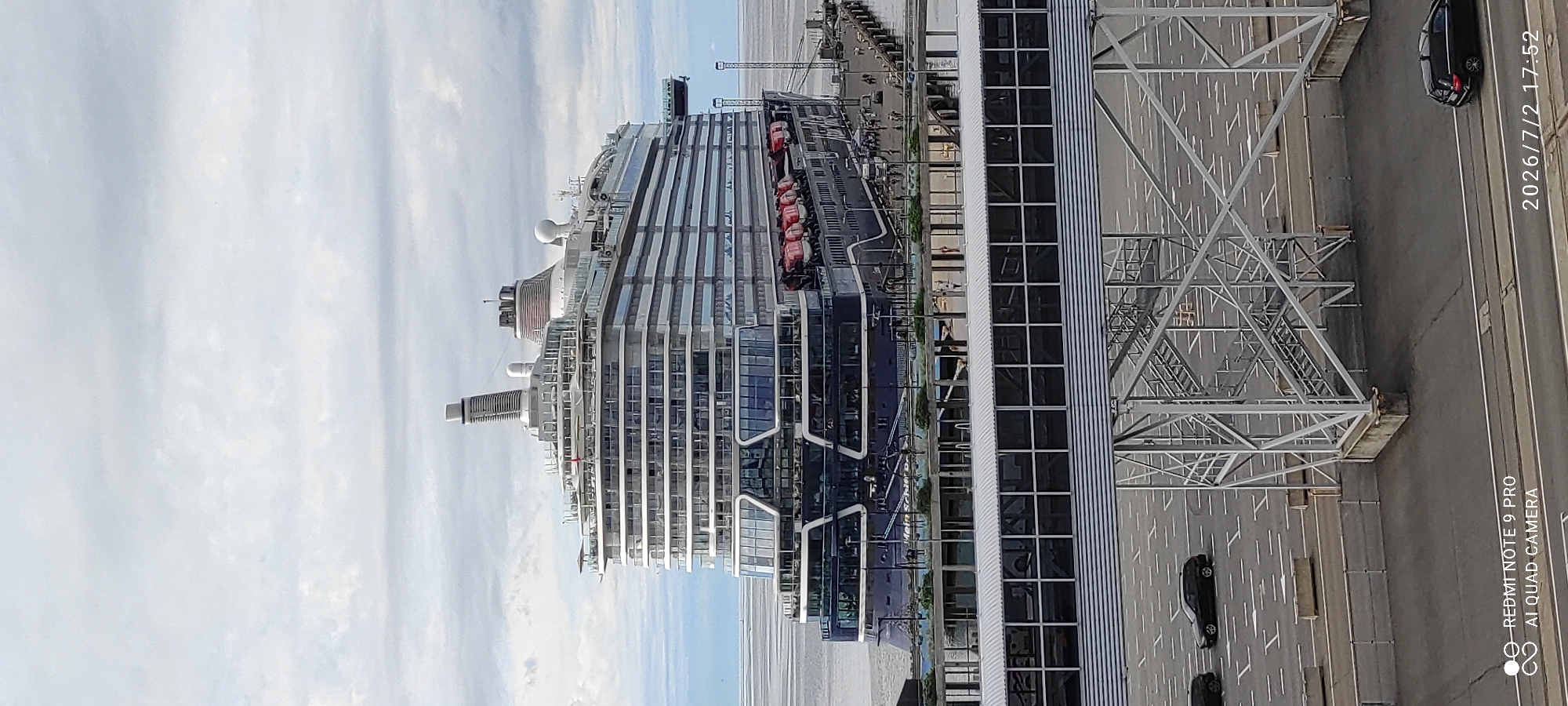 This is an aerial shot taken on a bright, overcast day, likely in the afternoon judging by the timestamp. The scene features a large, modern cruise ship docked alongside a substantial terminal building. The ship, with its distinctive sleek, multi-tiered design and numerous windows, is the primary subject. Life rafts are visible along its side.
Adjacent to the ship is a large, industrial-looking terminal structure with a metallic, angular design and many glass panels. The ground area around the ship and terminal is paved and marked with parking lines. A dark-colored car is visible on the pavement, suggesting a functional port or terminal environment.
The background consists of a hazy sky with clouds, and a calm body of water extending to the horizon. The location is confirmed as Tallinn, Estonia. There is visible text on the image: "2026/7/2 17:52" and "REDMI NOTE 9 PRO AI QUAD CAMERA". The overall impression is one of a busy, functional port with a focus on the impressive cruise liner.