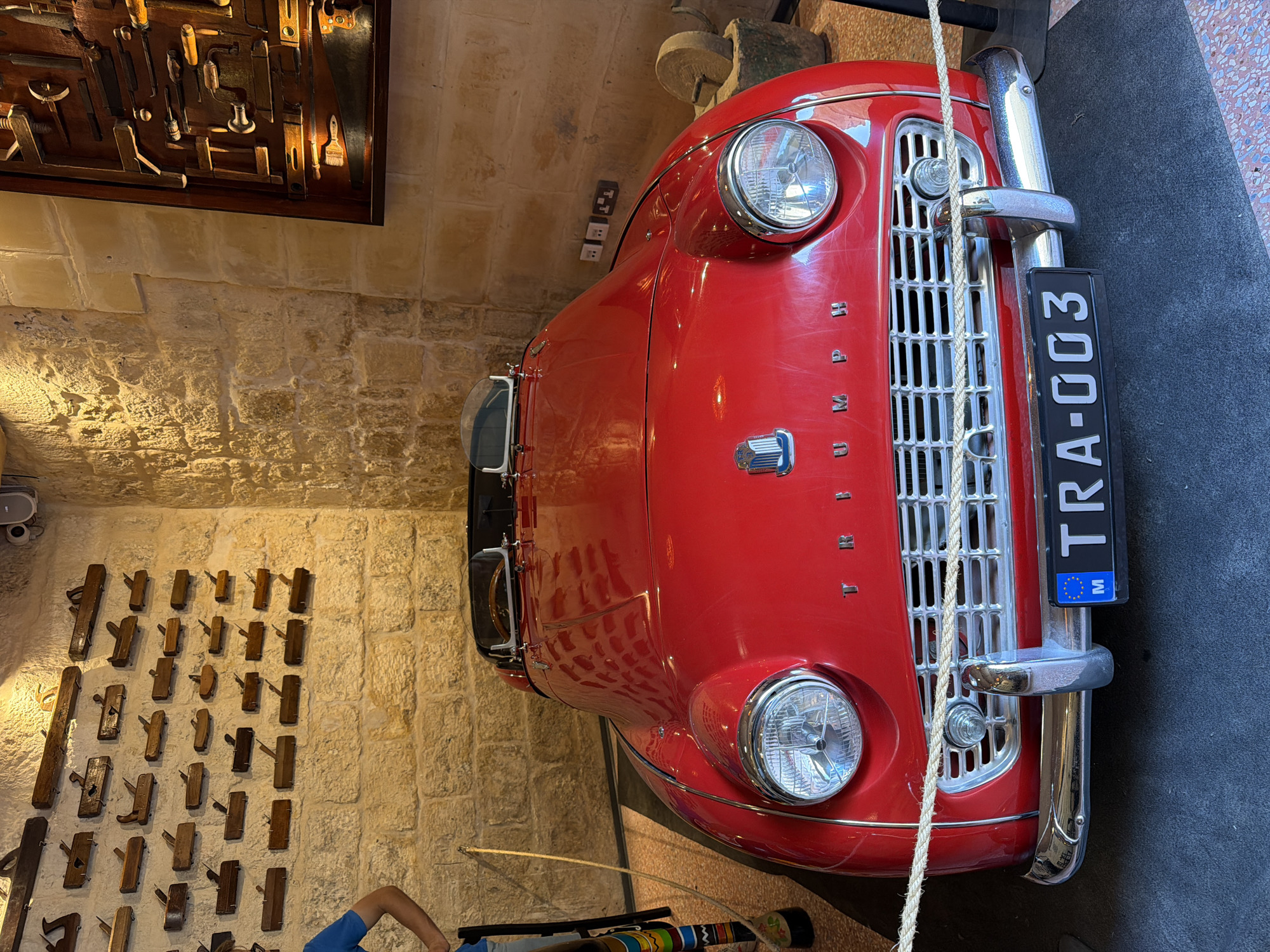 The image shows a red vintage Triumph car parked indoors, possibly in a museum or shop in Mdina, Malta. The car is the primary subject, with its front grille and headlights prominently displayed. The license plate reads "TRA-003" and features a European Union symbol.
The setting appears to be an old building with stone walls. To the left of the car, a collection of antique wooden tools, possibly woodworking tools, are mounted on the wall, adding to the historical ambiance. A glimpse of a person wearing a blue shirt is visible in the lower part of the image, suggesting there are visitors or staff present. The lighting is warm, possibly indicating indoor lighting, and there are no clear indications of the time of day or weather outside. The overall impression is one of a preserved historical display, showcasing a classic car and artisanal tools.