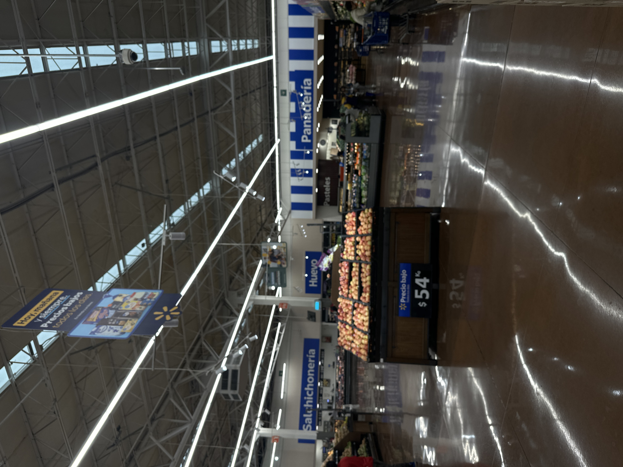 The image depicts the interior of a Walmart store, specifically within the location in the city of Jesús del Monte, Mexico. The scene is oriented with the aisle running away from the viewer. Overhanging signs designate different sections, including "Panaderia" (Bakery), "Salchichoneria" (Deli), "Huevo" (Eggs), and "Pasteles" (Pastries). Below "Huevo," displays of produce, likely apples, are visible, accompanied by a sign stating "$54" with "Precio bajo" (Low Price). A sign promoting "Hoy, mañana Y siempre. Precios bajos todos los dias" (Today, tomorrow, and always. Low prices every day.) is visible at the aisle entrance. People are present throughout the aisle. The environment consists of fluorescent lighting fixtures in the ceiling above shelving units with produce displays. The floor is shiny.