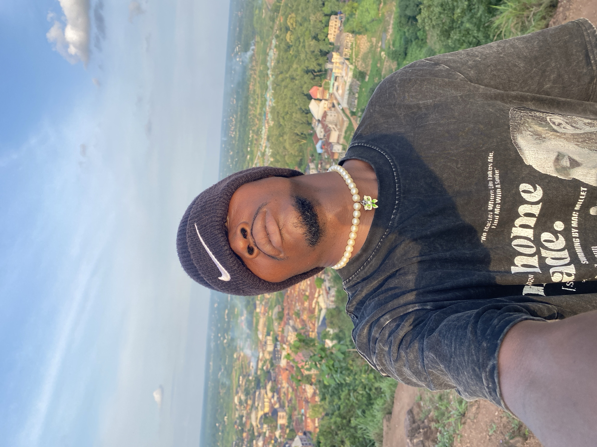 A man in a black beanie and a dark t-shirt stands on a hillside overlooking the city of Abakaliki, Nigeria. The man is wearing a pearl necklace with a green butterfly pendant. The sky is blue with some clouds. The city below is a mix of buildings and trees. The man is looking up at the sky, with his face tilted back. He is holding the camera in his left hand, taking a selfie.