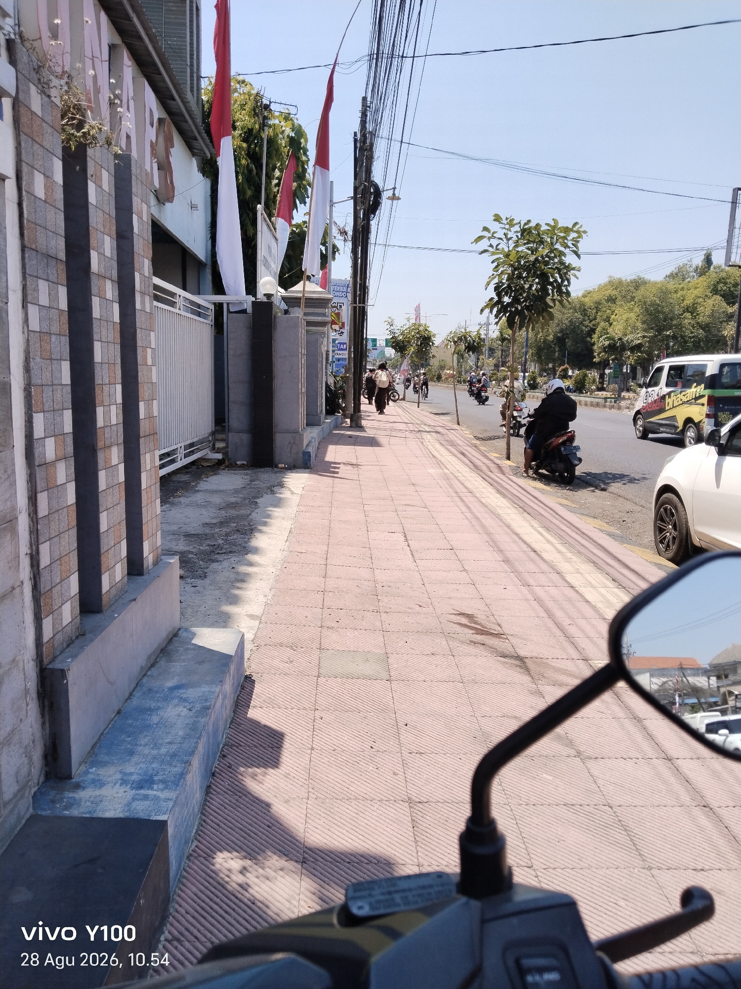 The image depicts a street scene in Situbondo, Indonesia. The perspective is from the rider of a motorcycle, with a rearview mirror and handlebars visible in the foreground. A paved sidewalk with red tiles runs along the left side of the street, bordered by a patterned tiled wall of a building. Indonesian flags are prominently displayed, attached to poles along the sidewalk. The street itself is asphalted and shows moderate traffic, including cars and motorcycles. Several trees line the right side of the street, providing shade. The sky is clear and blue, indicating a sunny day. The overall impression is a typical urban street environment in Indonesia, characterized by local architecture, transportation, and national symbols.